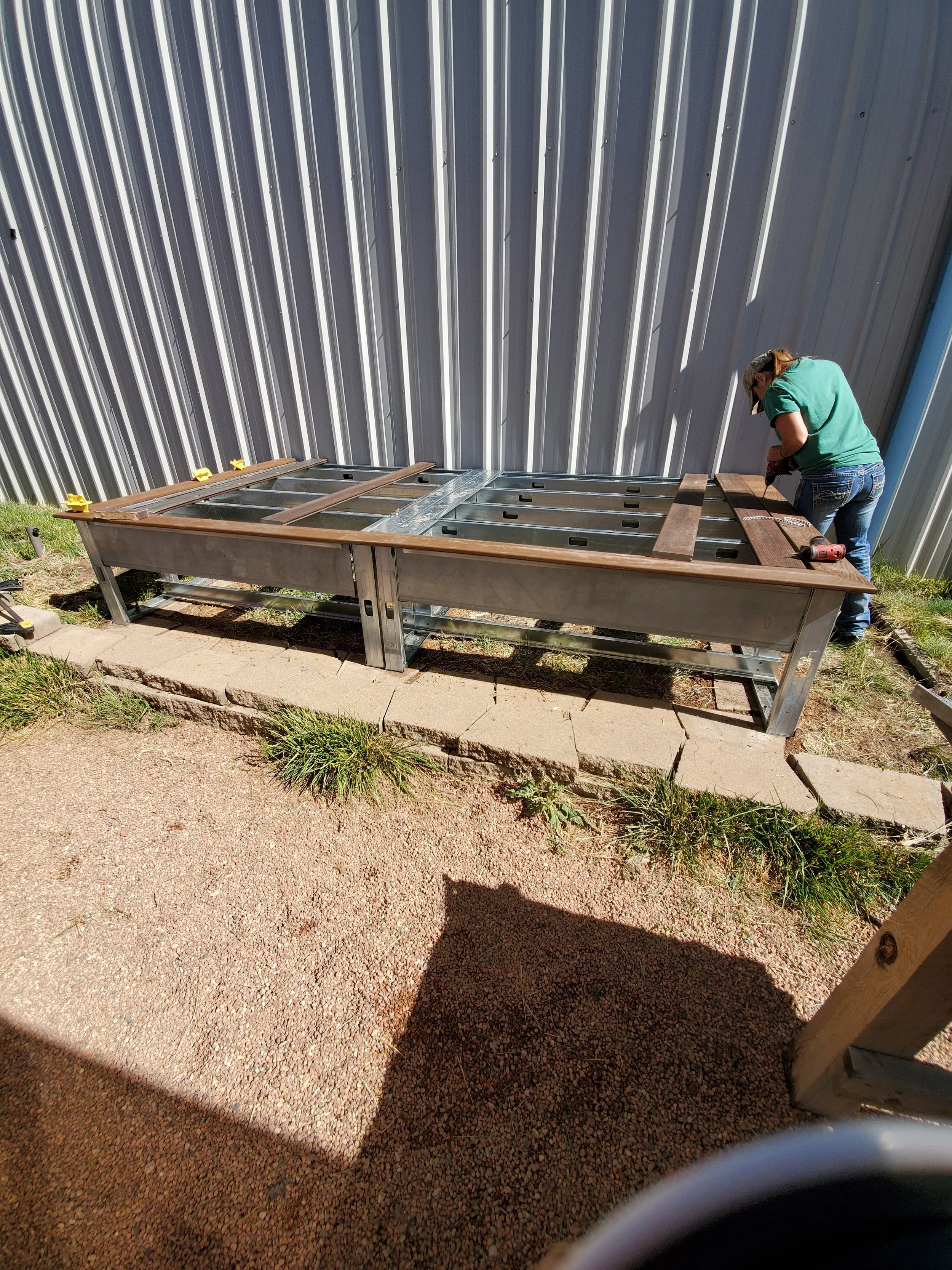 A woman is assembling a metal-framed outdoor grill or smoker on a paved patio next to a metal wall, with tools and wooden panels around her.