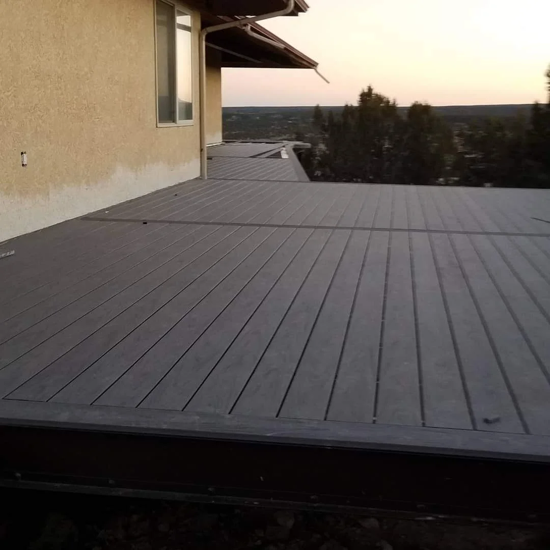 Empty rooftop deck with gray decking boards, beige exterior wall with a window, and trees in the distance at sunset.