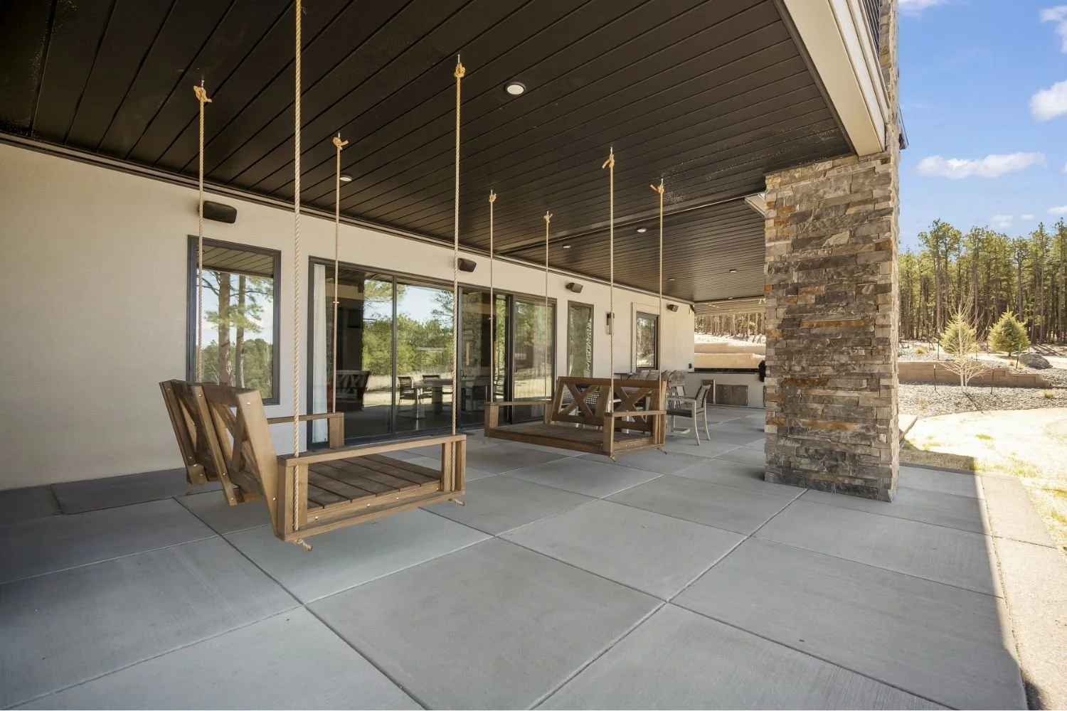 Covered patio with wooden swings hanging from the ceiling, a stone pillar, glass sliding doors, and outdoor furniture outside in a natural setting with trees.
