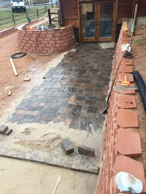 A brick walkway under construction leading to a building with double glass doors. The walkway has alternating finished brick sections and areas with sand and paving stones, with construction tools and materials around.