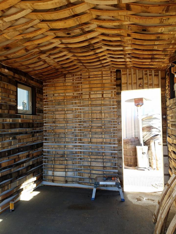 Interior of a small wooden structure under construction with curved wood ceiling, partially installed wall slats, a ladder, and a window.