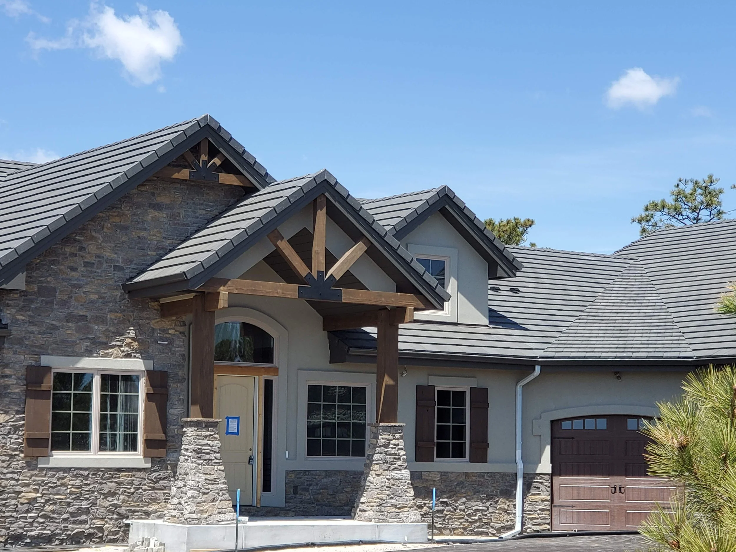 Front of a house with a stone and stucco exterior, multiple gable roofs with gray shingles, and a front yard with plants, under a blue sky with a few clouds.