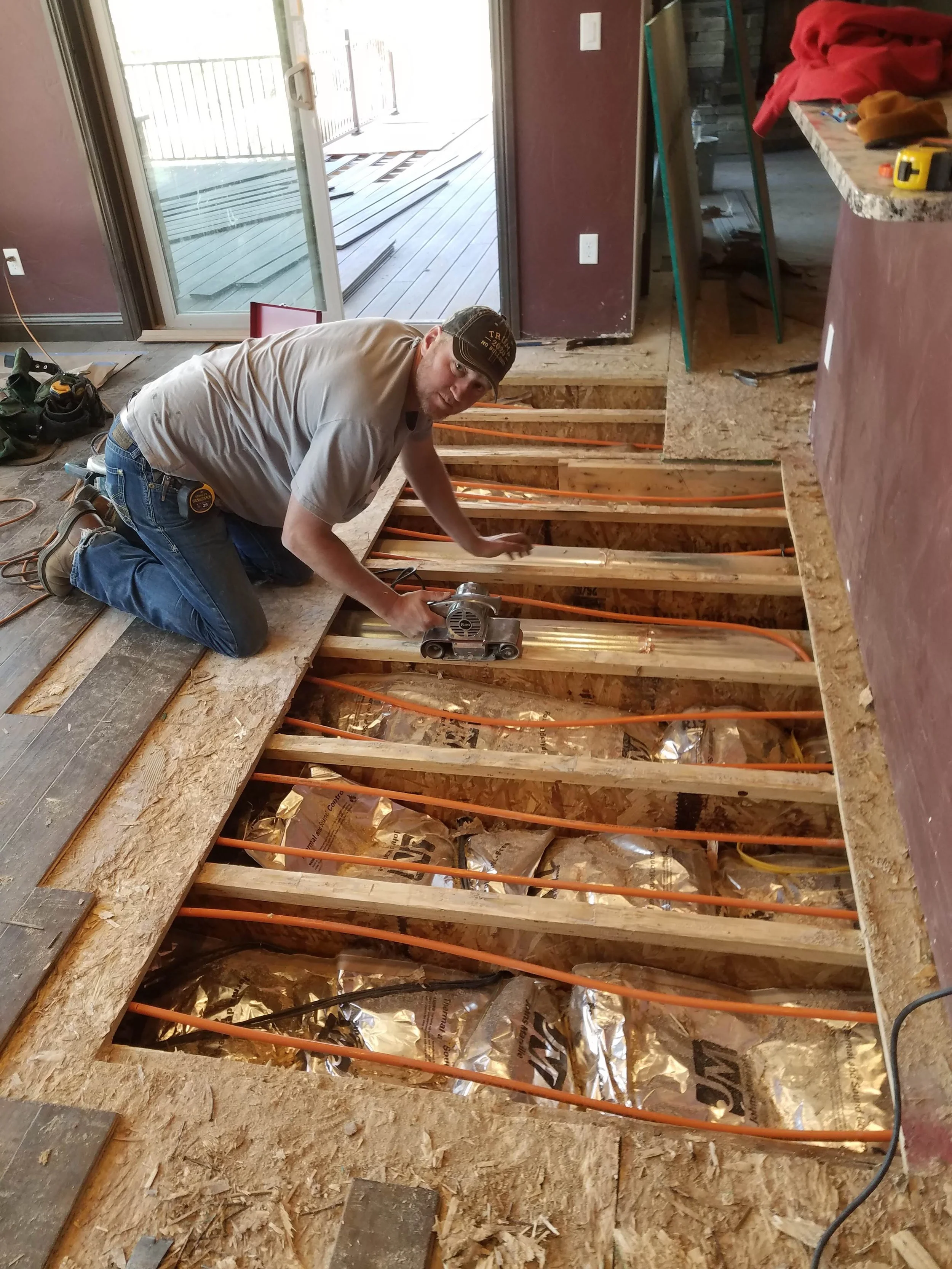 A man kneels on the floor of a room under renovation, using a power tool on wooden flooring. The floor is partially removed, exposing ground and orange wiring. There is a sliding glass door leading to an outdoor deck area with wooden planks. Construc