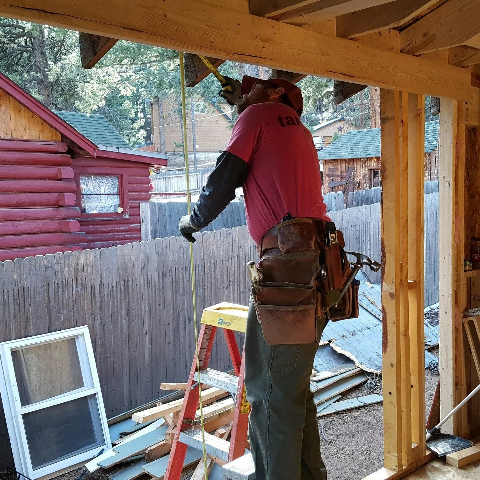 A construction worker standing on a ladder measuring a wooden ceiling beam with a tape measure at a building site.
