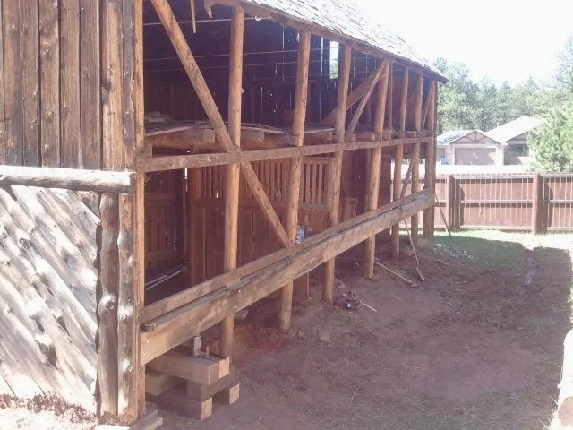 Wooden structure under construction, elevated on wooden supports, with a roof and part of a fence visible in the background.