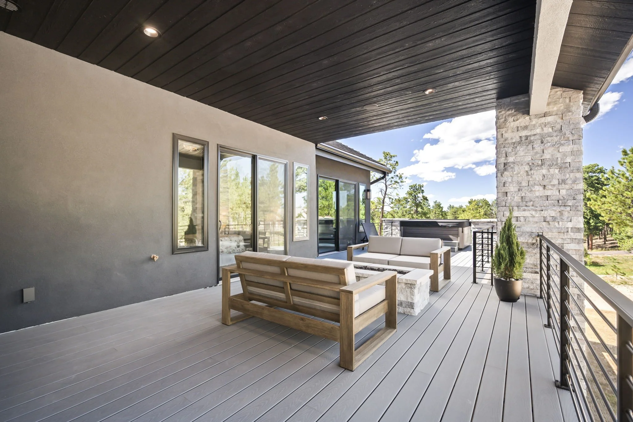 A modern outdoor balcony area with wooden floorboards, black metal railing, and outdoor furniture including a wooden bench and white cushioned seating. There's a potted plant, large glass doors leading to indoor space, a stone pillar, and a dark wood