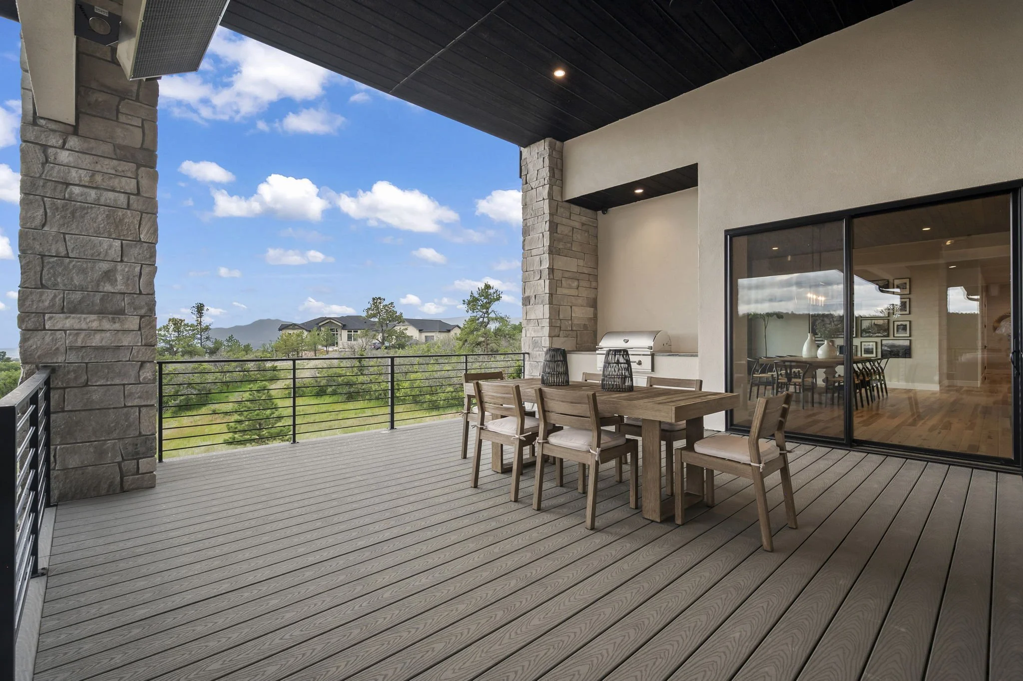 A spacious outdoor balcony with wooden flooring, a dining table with chairs, stone pillars, sliding glass doors, and a view of trees, houses, and mountains under a partly cloudy sky.