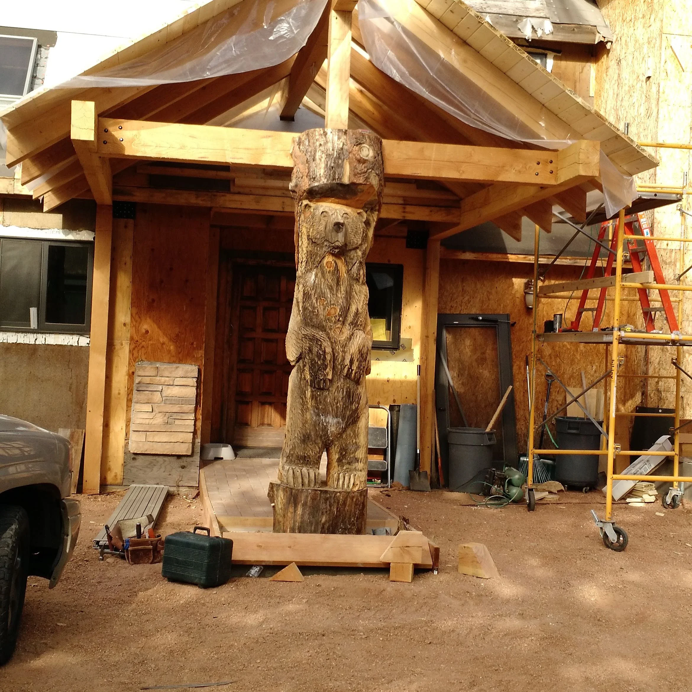 A wooden house under construction with a tree trunk carved into a bear's face supporting the roof. Building materials and tools are scattered around, and there is scaffolding on the right side.