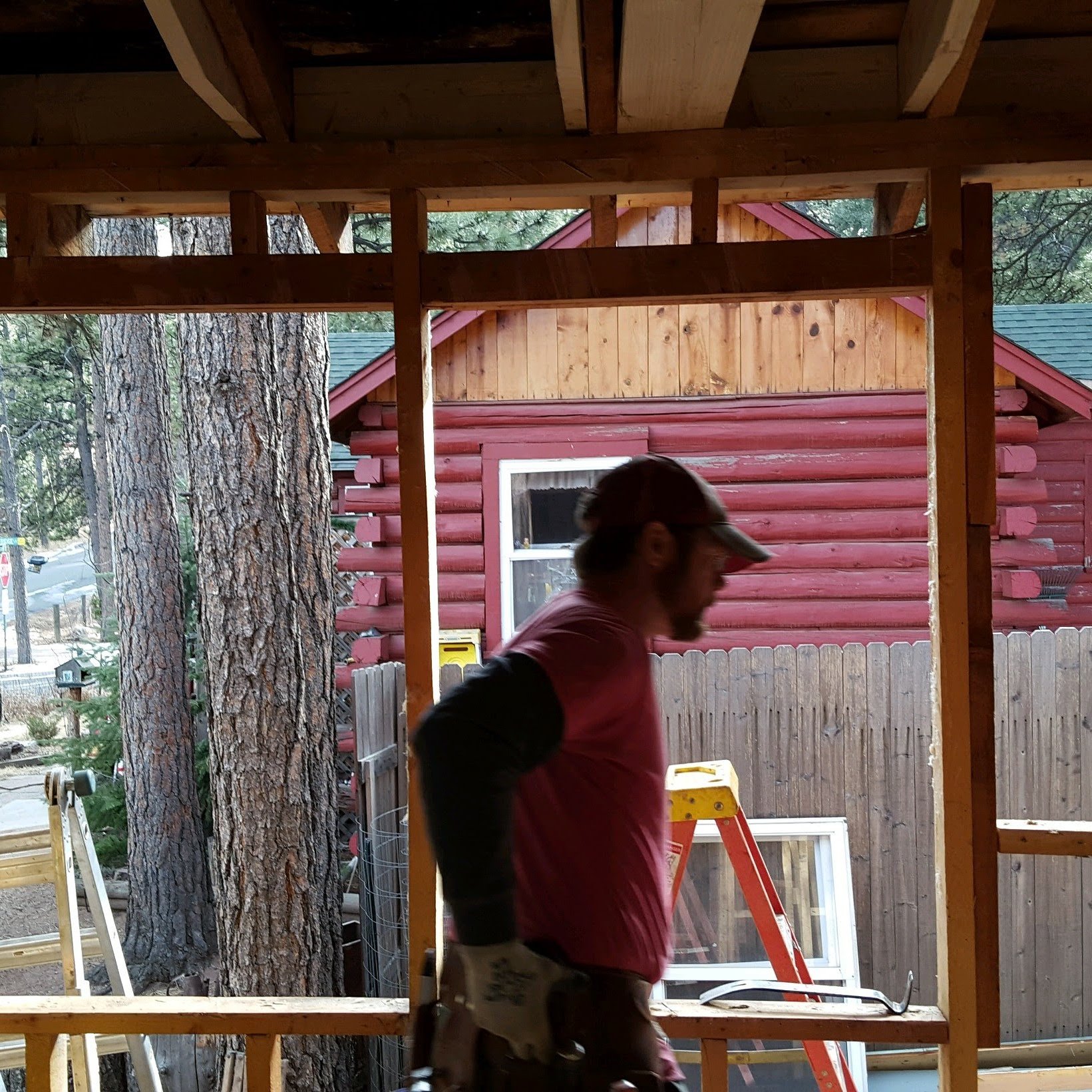 A person wearing a cap working on a wooden construction framework outdoors, with trees and a red wooden house in the background.