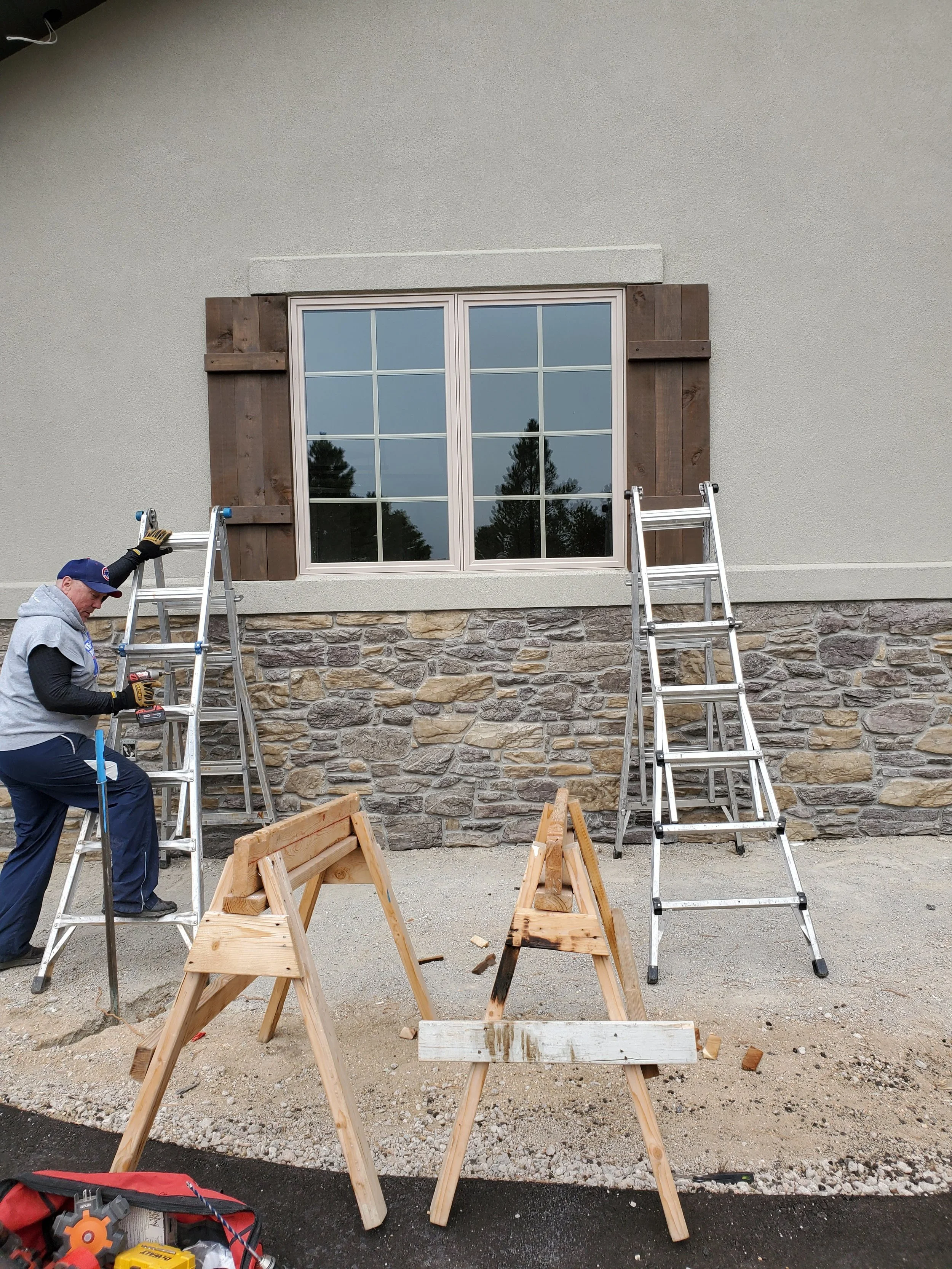 Person working on scaffolding outside a house finishing installation of Cedar shutters with a large window, stone foundation, and unfinished wooden window shutters, with construction tools and woodwork on the ground.