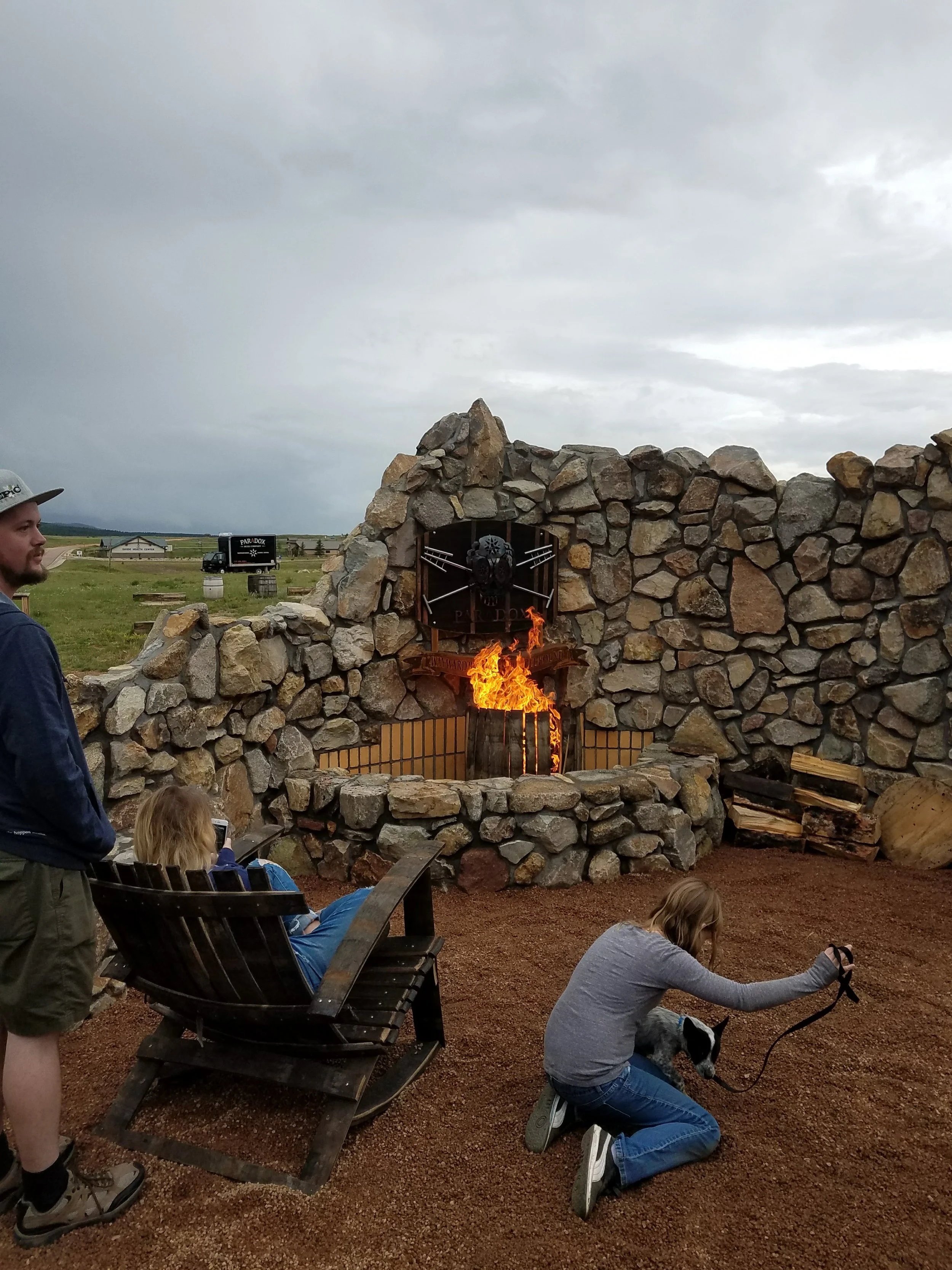 A group of people, including a man, a woman, and children, gathered outdoors near a stone fireplace with a skull and crossed swords emblem, on a cloudy day.