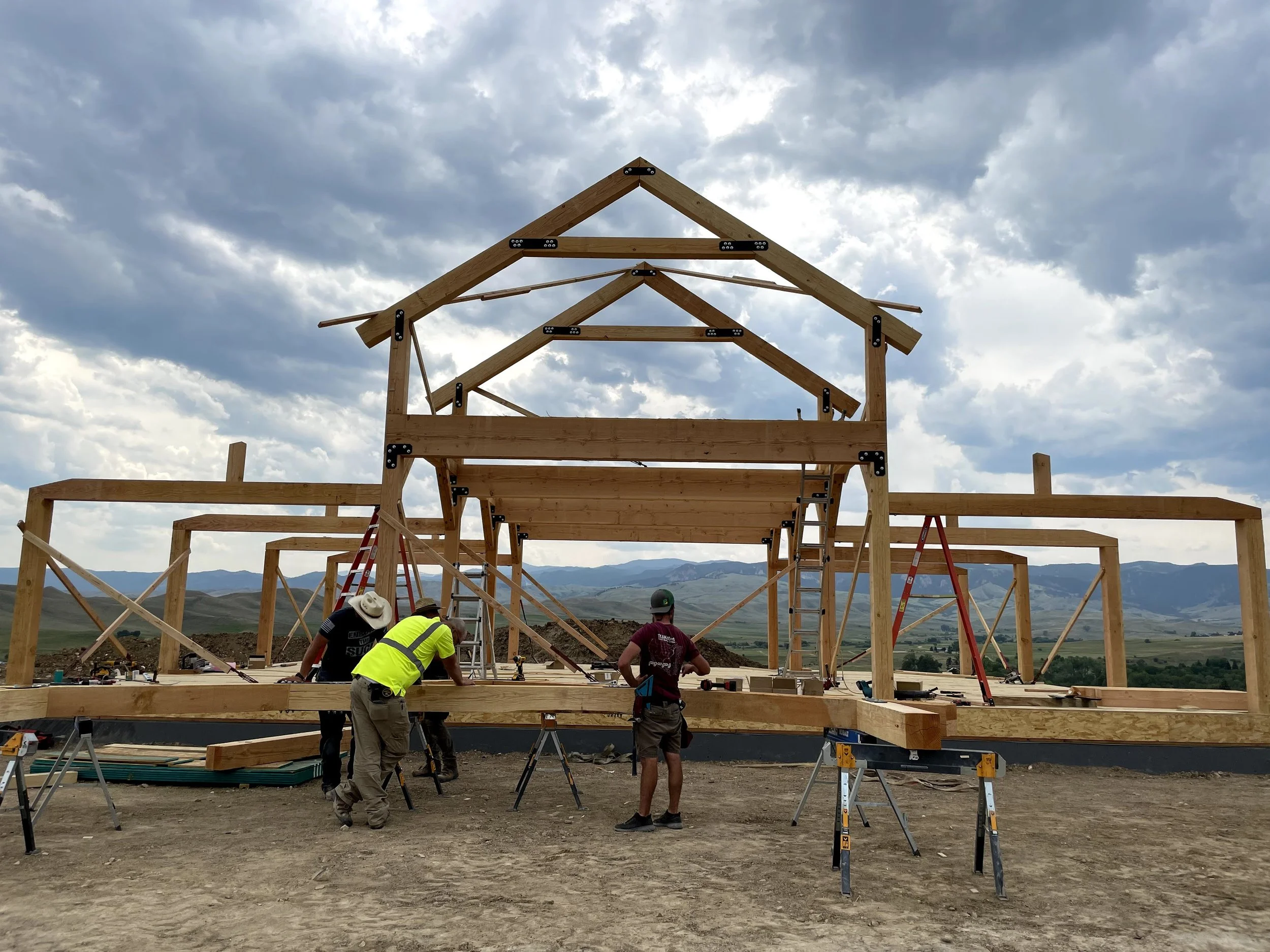 Construction workers building a Post and Beam house with a gabled roof frame against a cloudy sky and distant mountains near Sheridan Wyoming