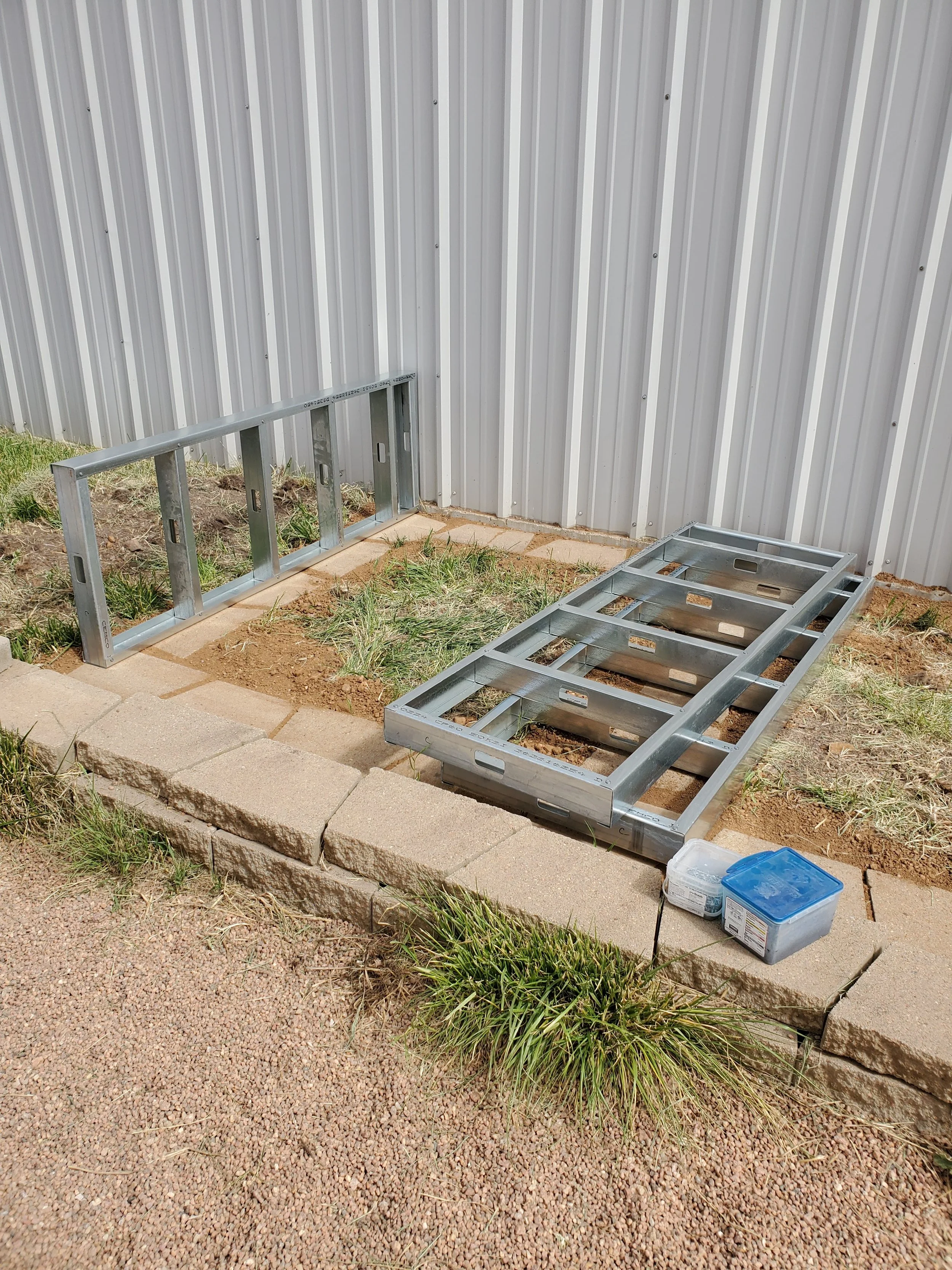 Partially assembled metal frame of a small structure on a brick and dirt foundation, with a corrugated metal wall in the background and construction supplies nearby.