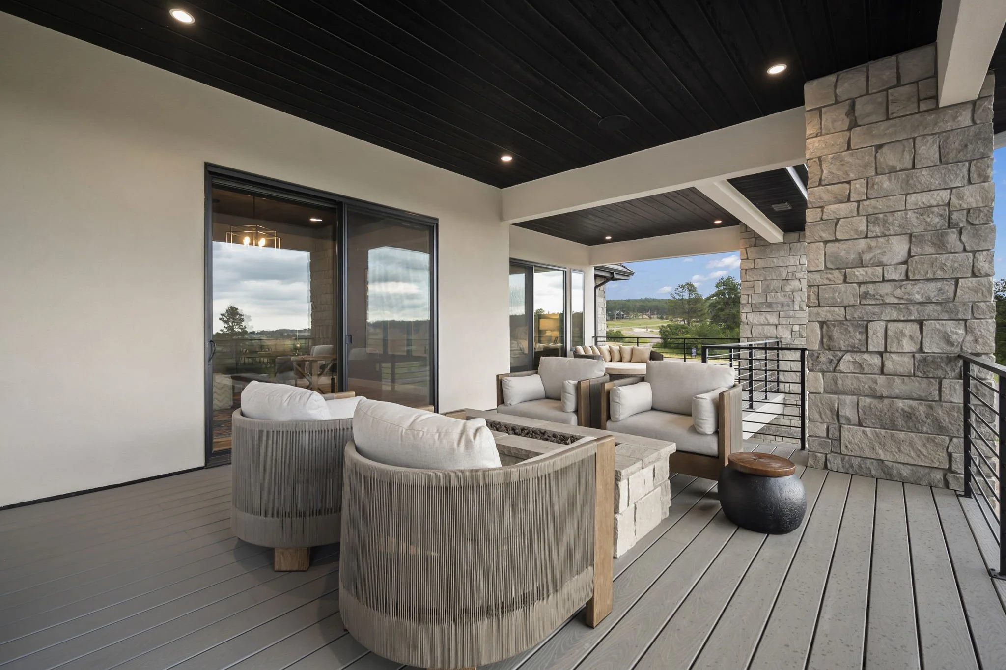 A modern outdoor balcony with beige cushioned chairs and a stone pillar, overlooking a landscape with trees and a lawn.
