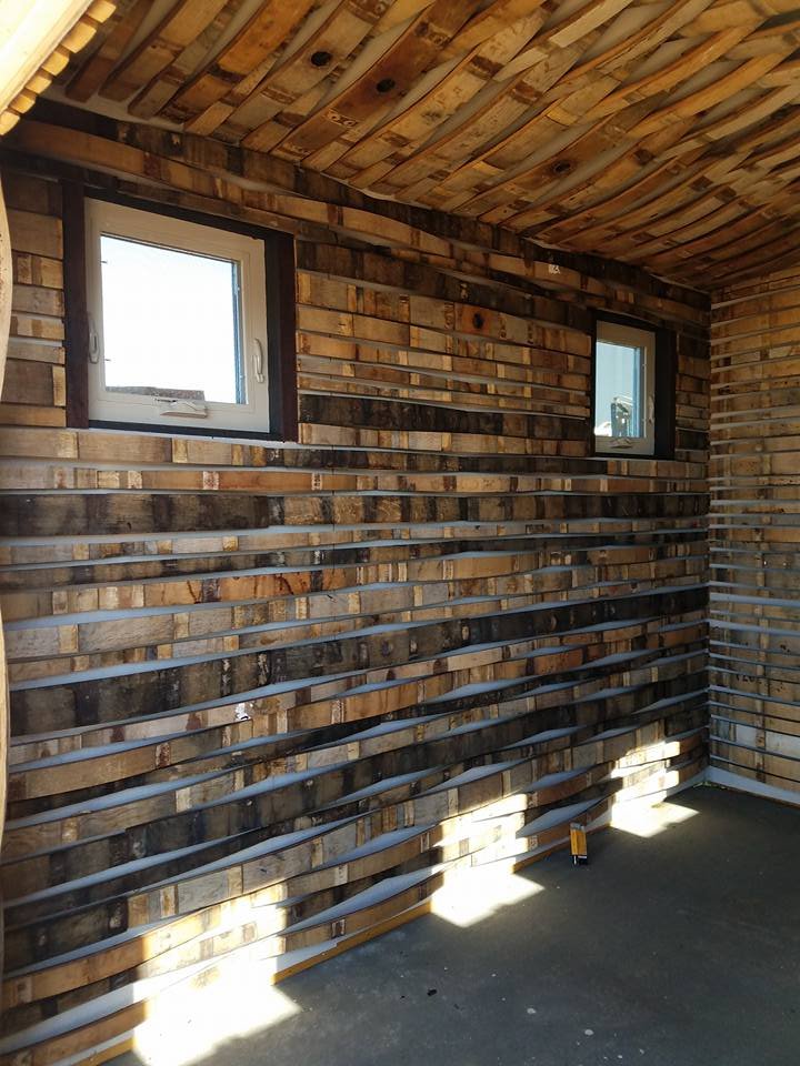 Interior of a room with wooden slat walls and ceiling, two small windows, with sunlight casting shadows on the floor.
