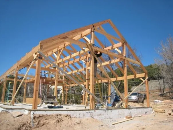 A timber frame house under construction on a dirt lot with a clear blue sky in the background.