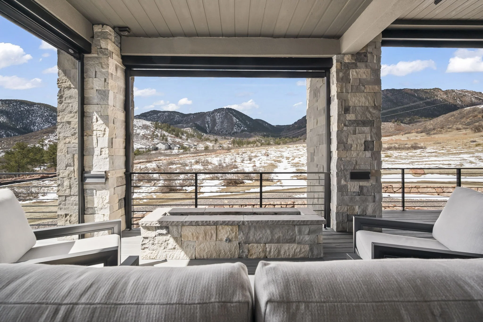 View from a covered patio area with white cushioned seating and a stone fireplace, overlooking a snowy landscape with mountains and a partly cloudy sky.