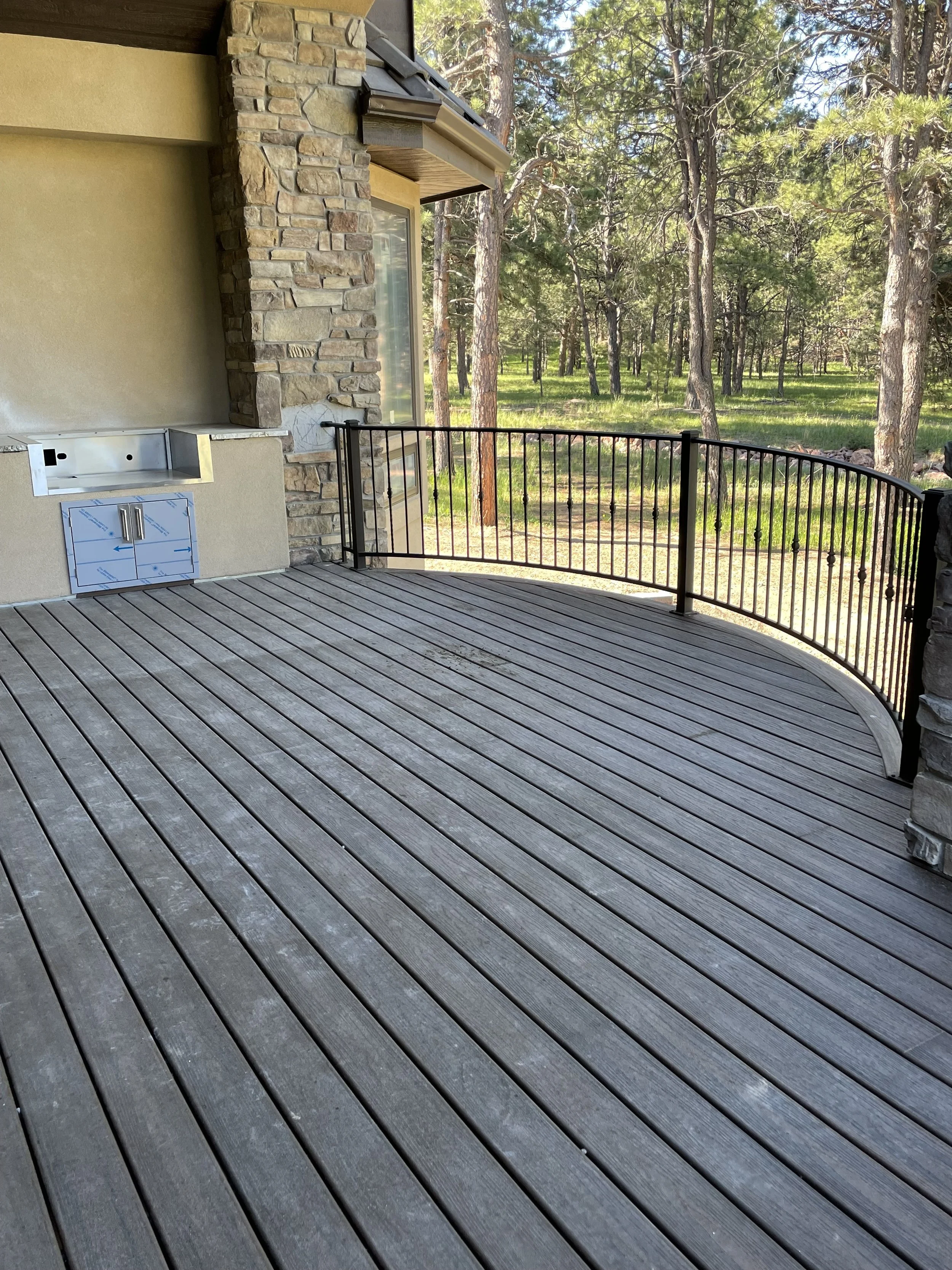 View of a wooden deck with black metal railing, part of a house with stone and stucco exterior, surrounded by trees and forest.