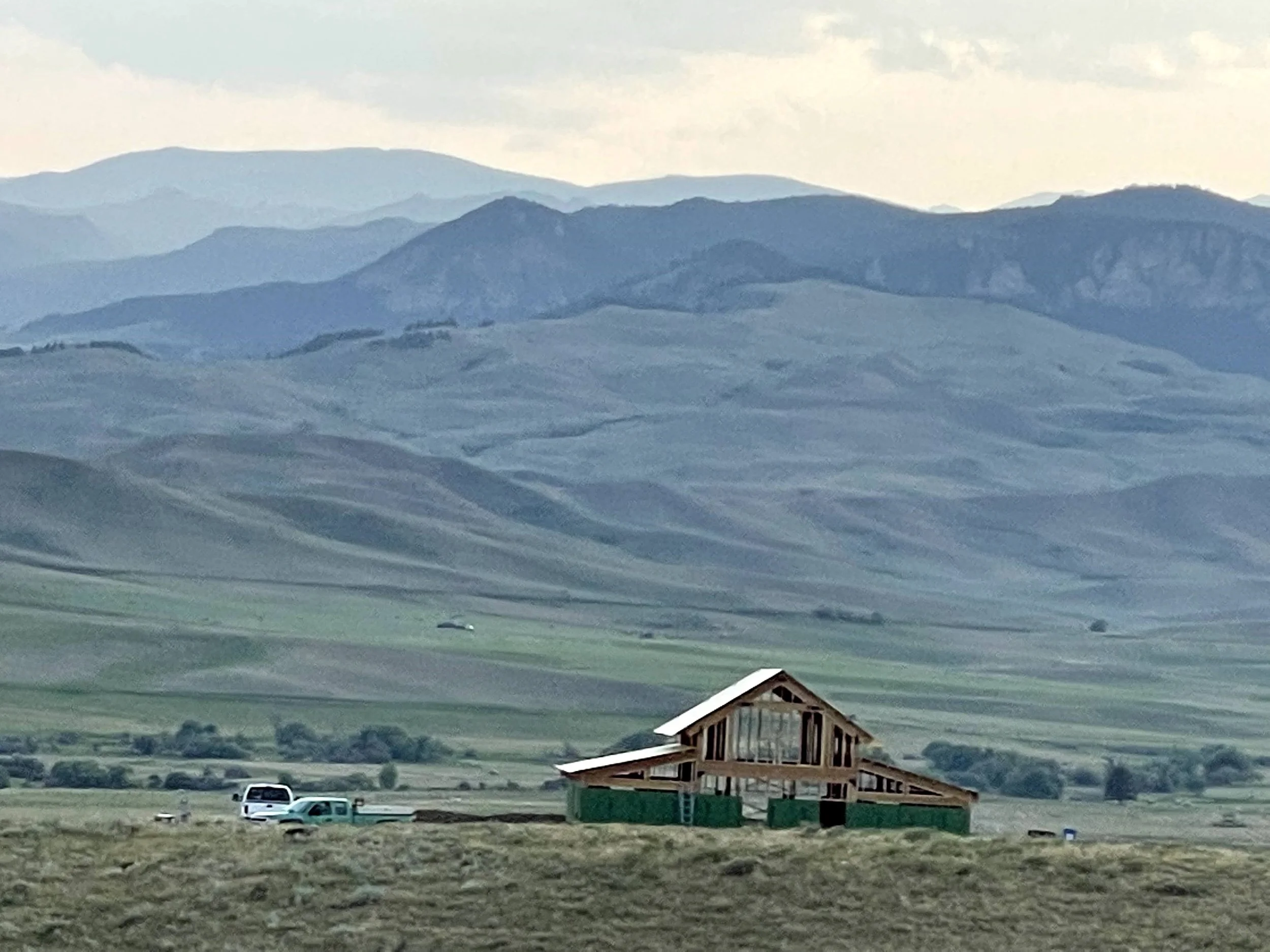 A rural landscape with mountains in the background, a barn in the foreground, and a few cars parked on the grass in front of the barn.