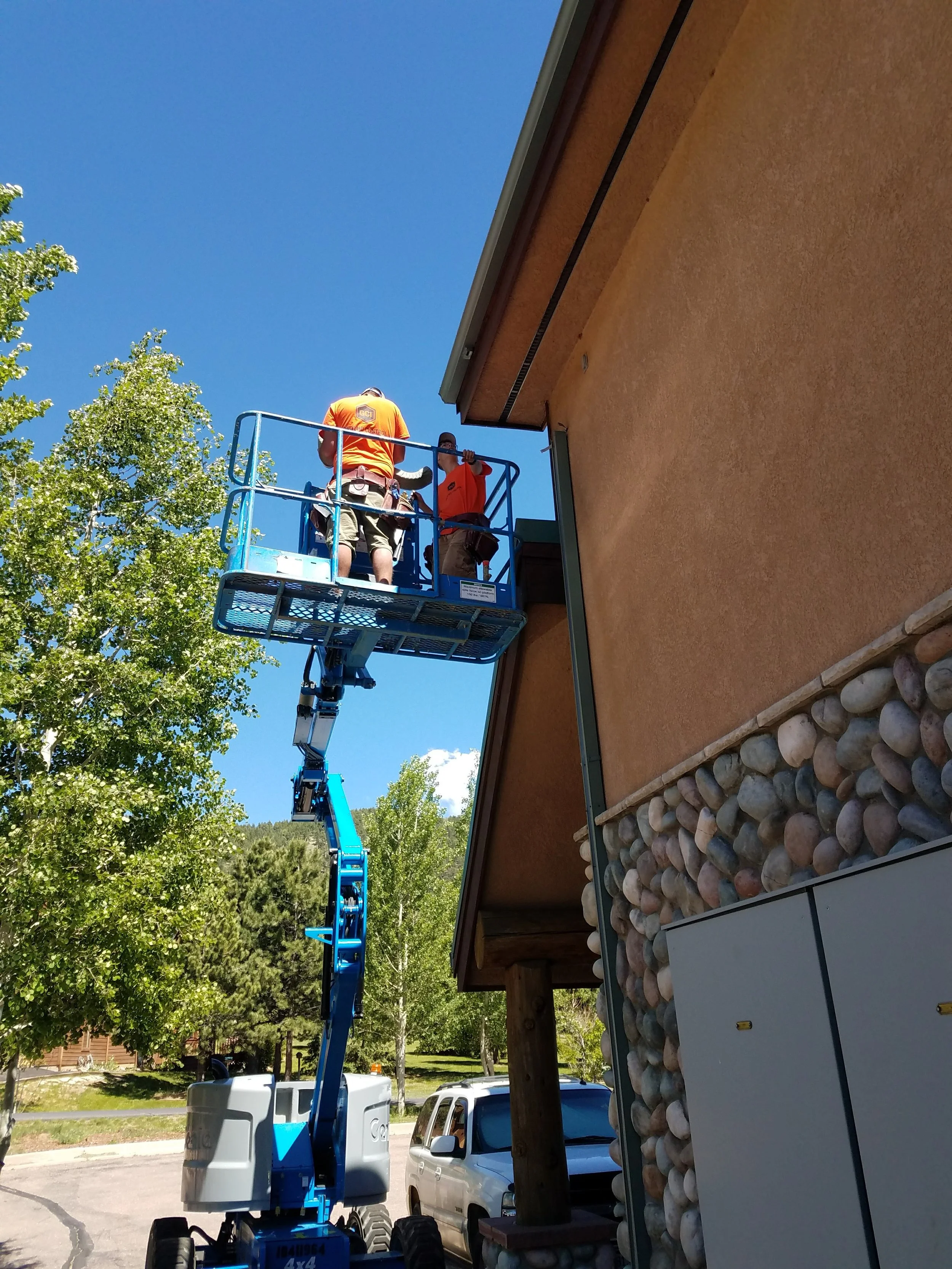 Two workers standing on a blue cherry picker elevated near the side of a building, with one facing away from the camera and the other facing towards it, working on the building's exterior during daytime with a clear blue sky overhead.