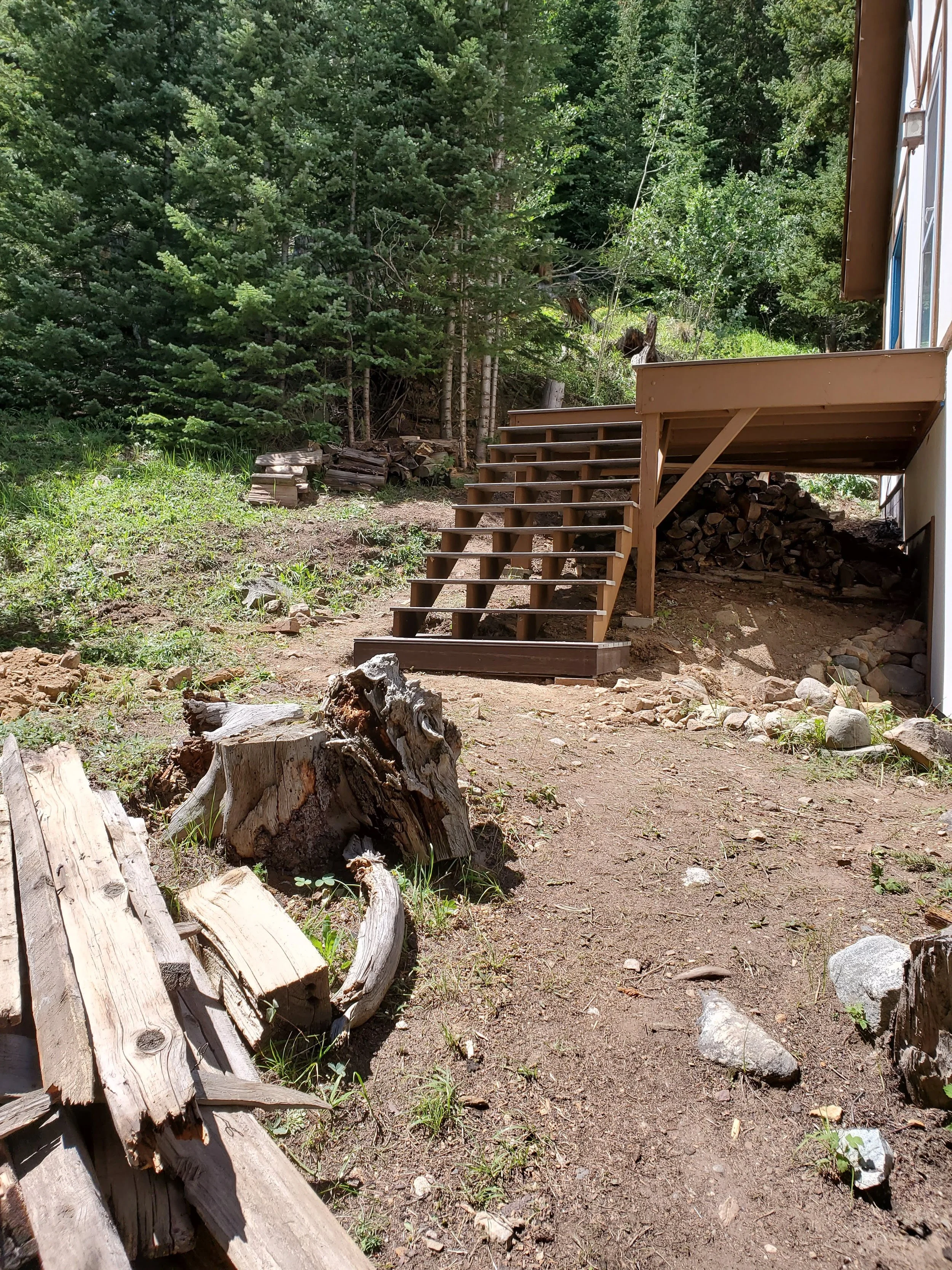 Wooden stairs leading down to a dirt path surrounded by greenery and trees, with a pile of firewood under the deck.