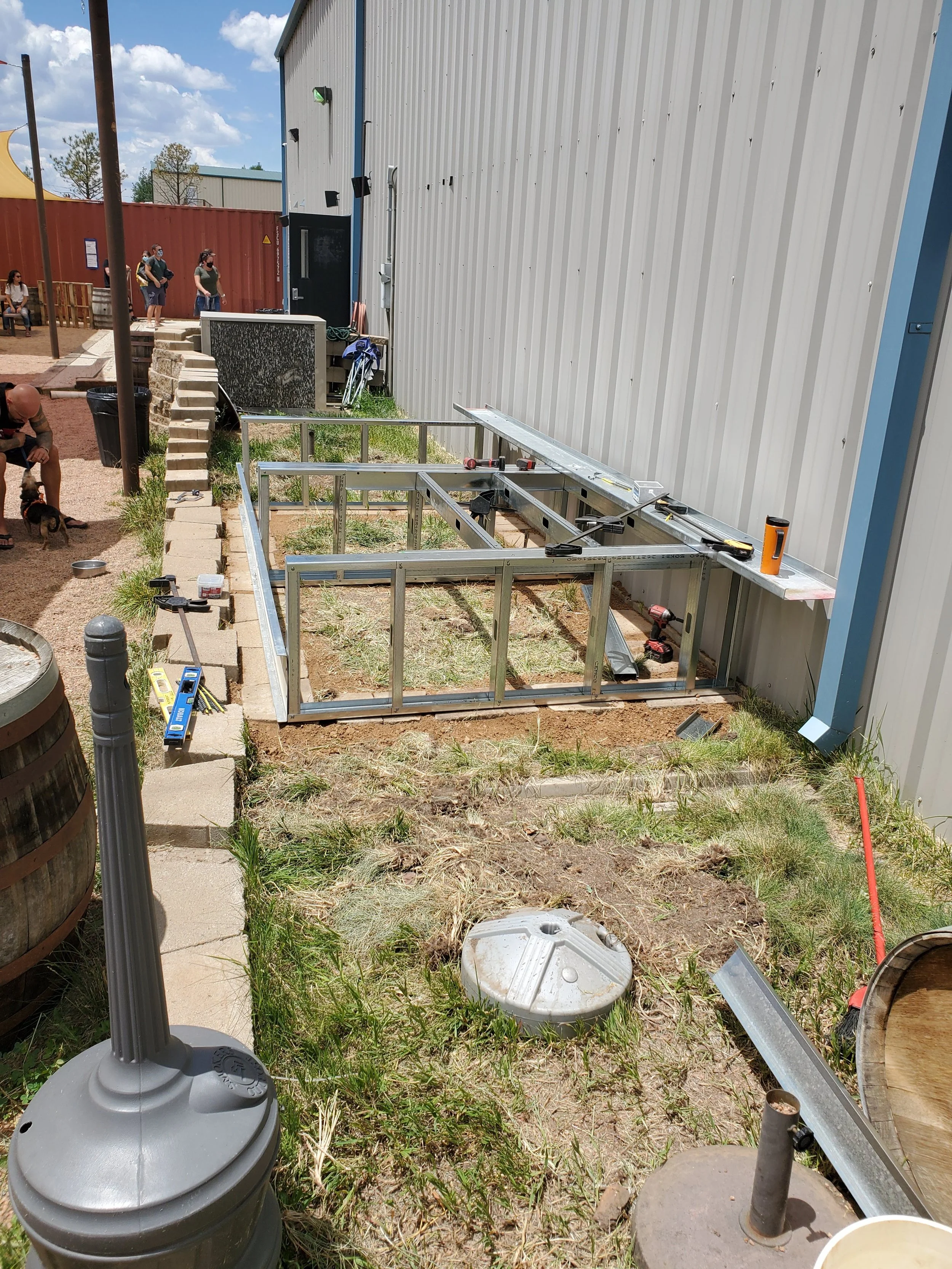 Construction site with metal framework for an outdoor structure next to a building. Tools and materials are scattered around, including a level, screws, and metal beams. A large grey umbrella is in the foreground, and people are waiting in line in th