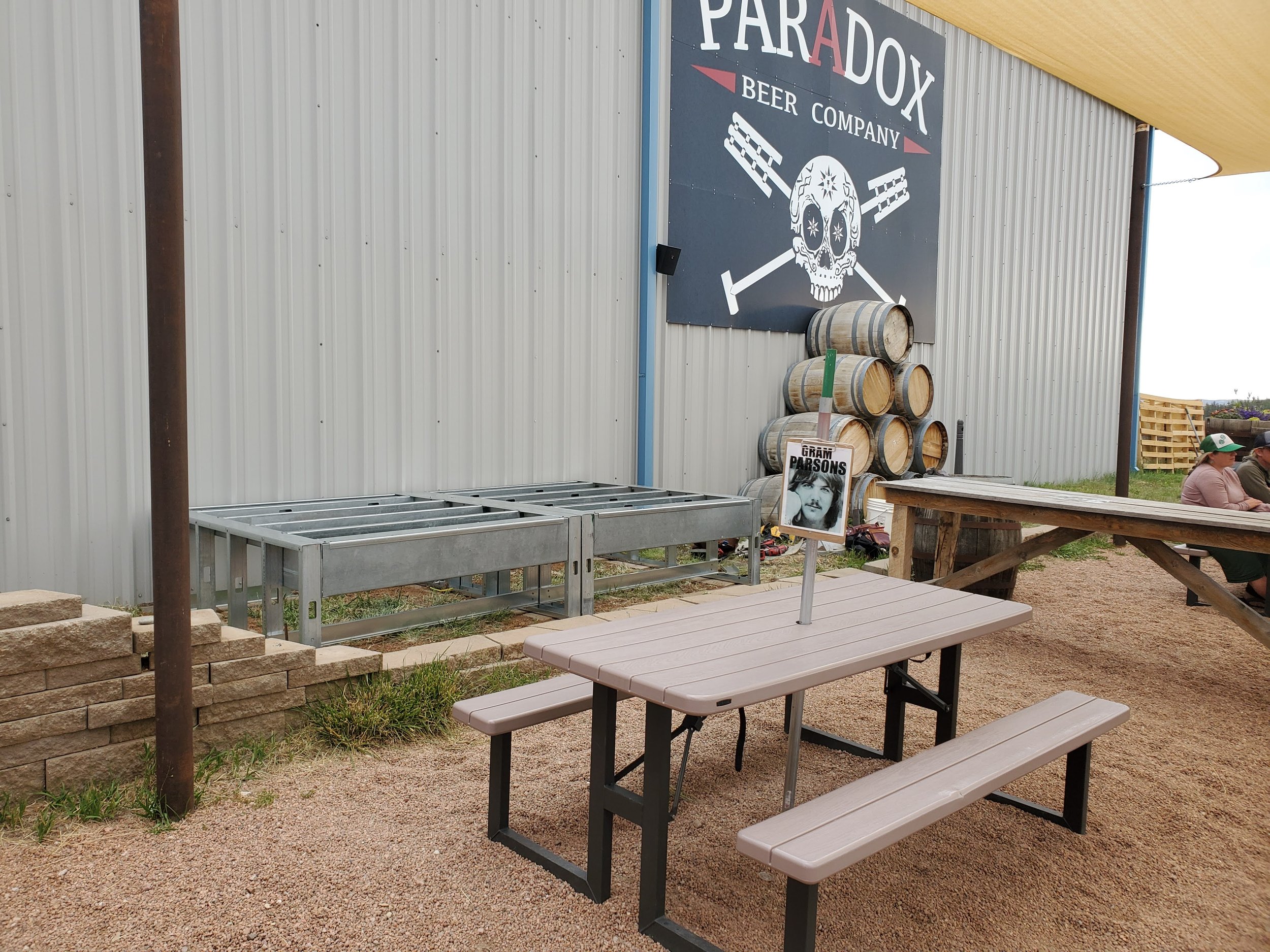 Outdoor seating area with picnic tables, stacked barrels, and a large sign for ParaDx Beer Company on the building wall with skull and crossbones graphic.