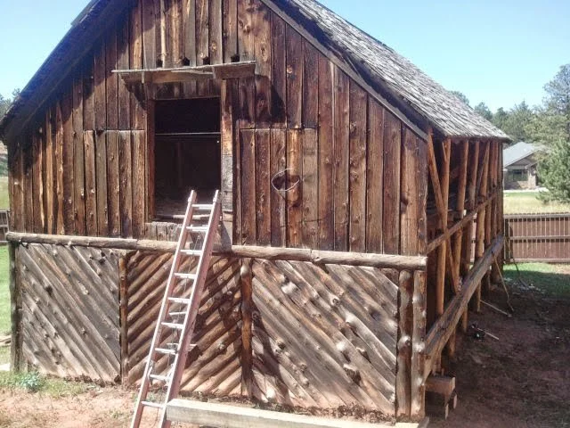 Wooden barn with a ladder leaning against the front, open window at the top, and a sloped roof, in a grassy outdoor area.