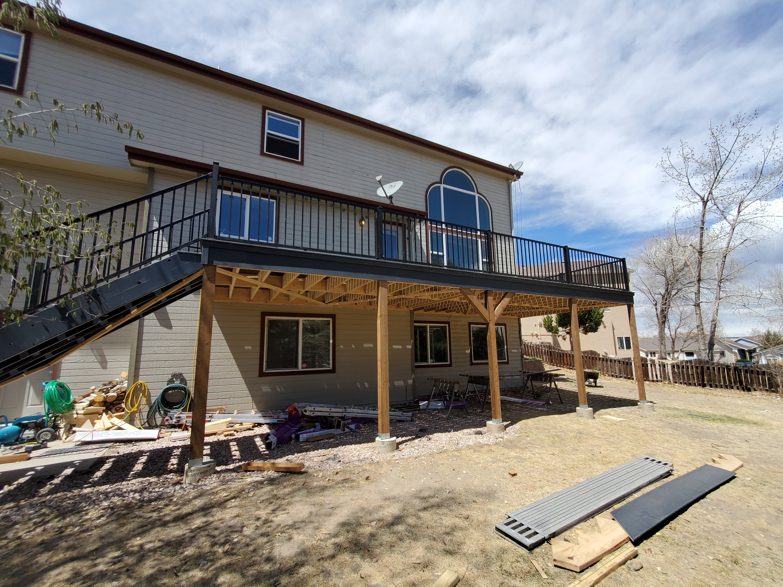 Backyard under construction with elevated deck being built, tools, and materials on the ground, and trees in the background.