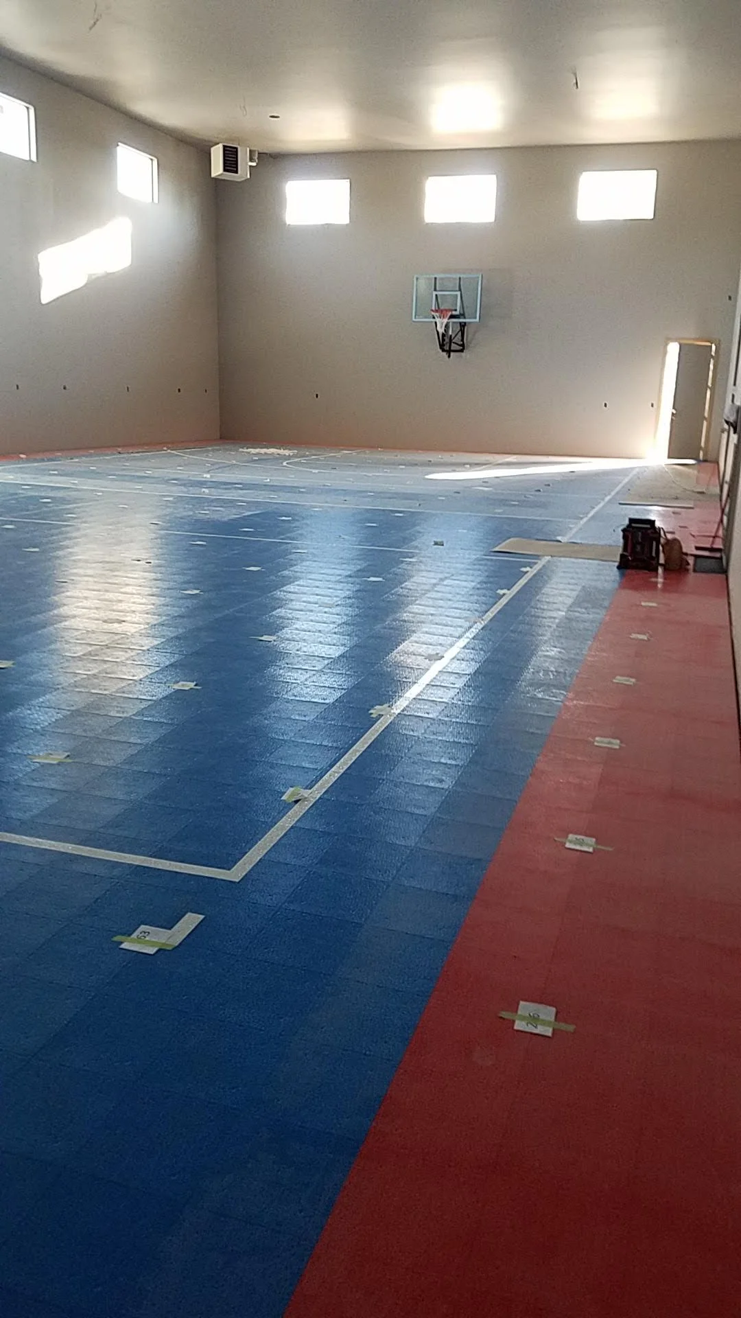 An indoor basketball court with blue and red flooring, and a basketball hoop on the wall. The court is under construction or renovation, with numbered tape strips on the floor.