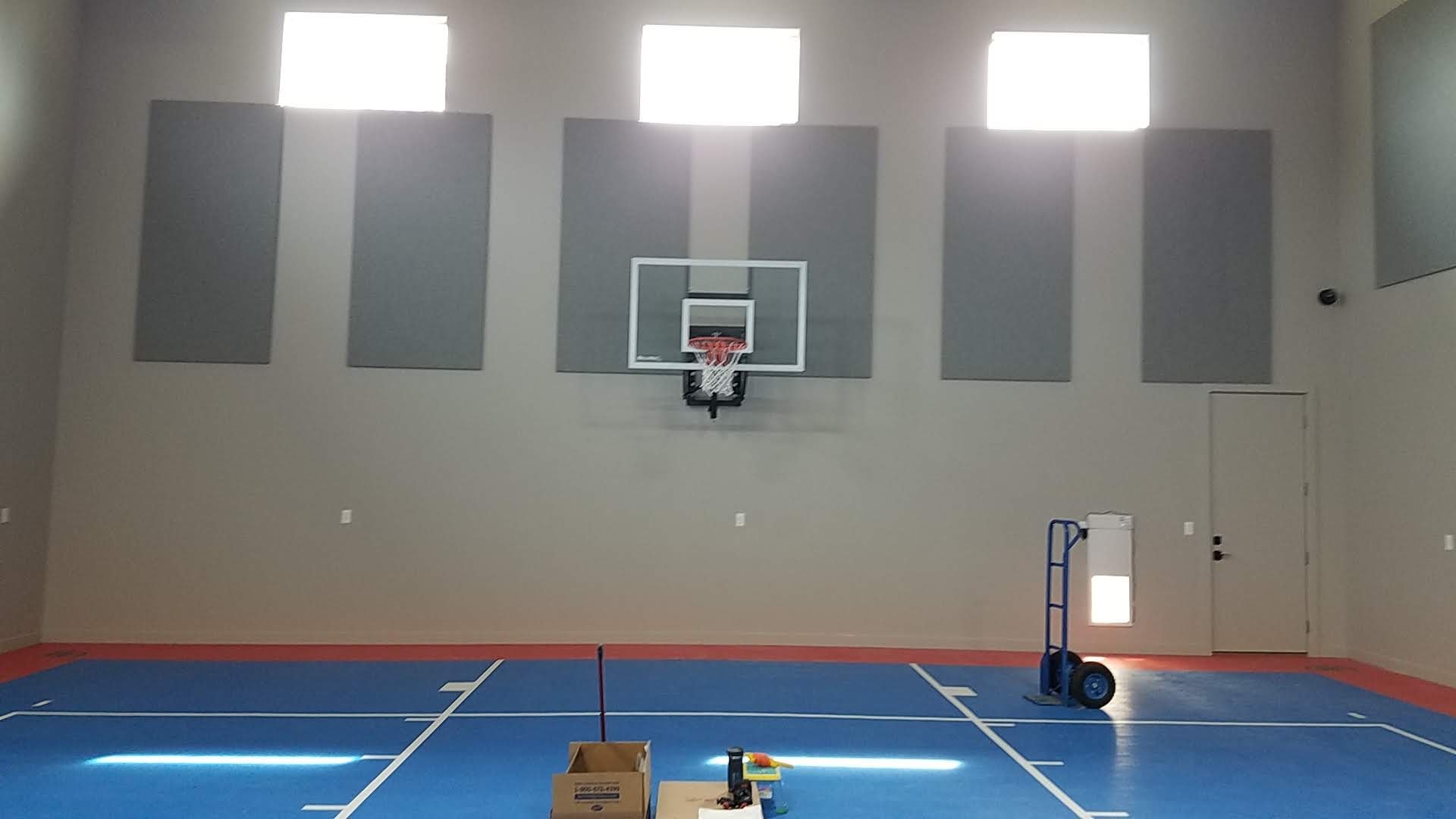 Indoor gym with blue court, basketball hoop, gray sound panels on the wall, high windows, and equipment including a dolly and cardboard box.