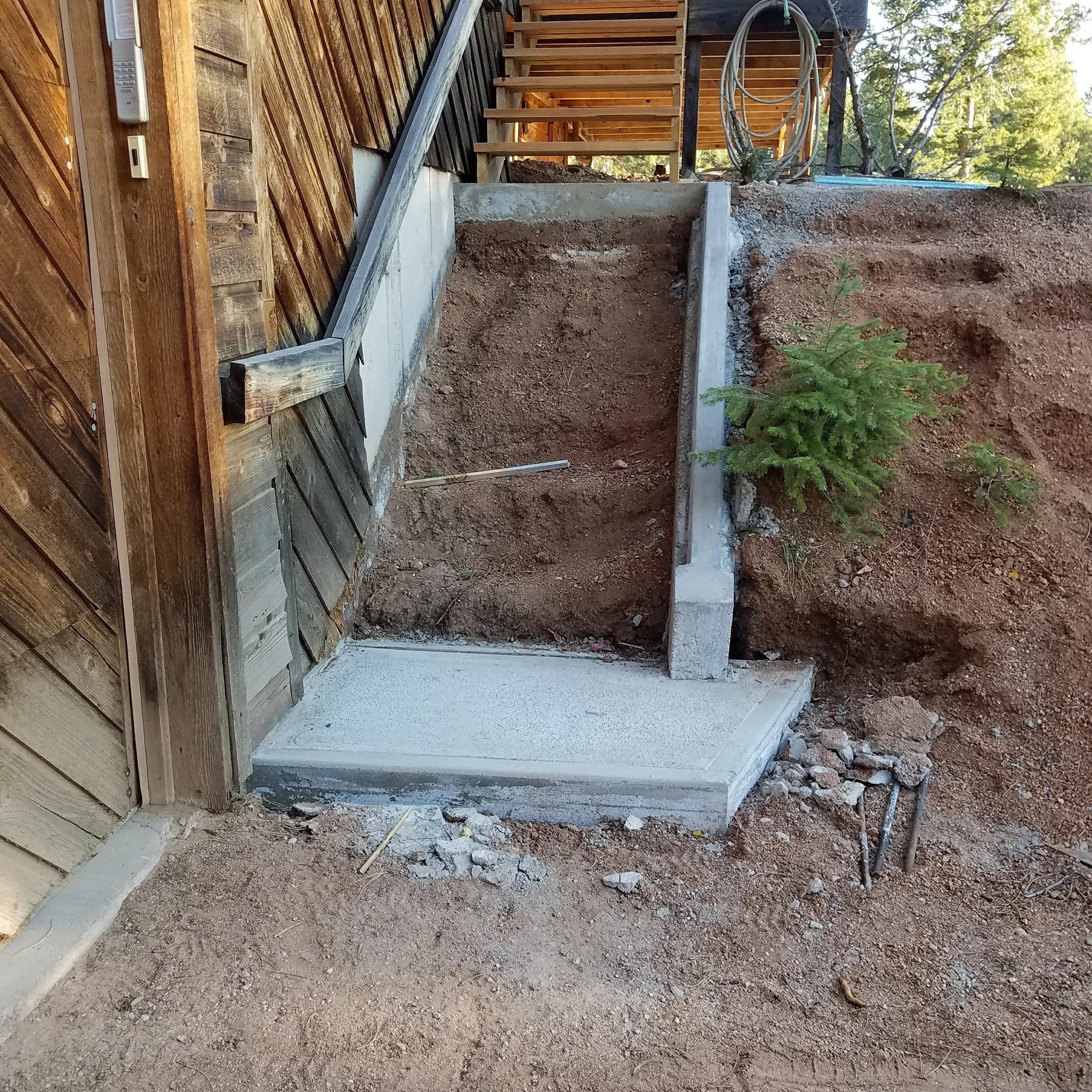 Construction site with a concrete foundation, dirt, small plants, and a wooden staircase leading to an elevated area, with trees in the background.