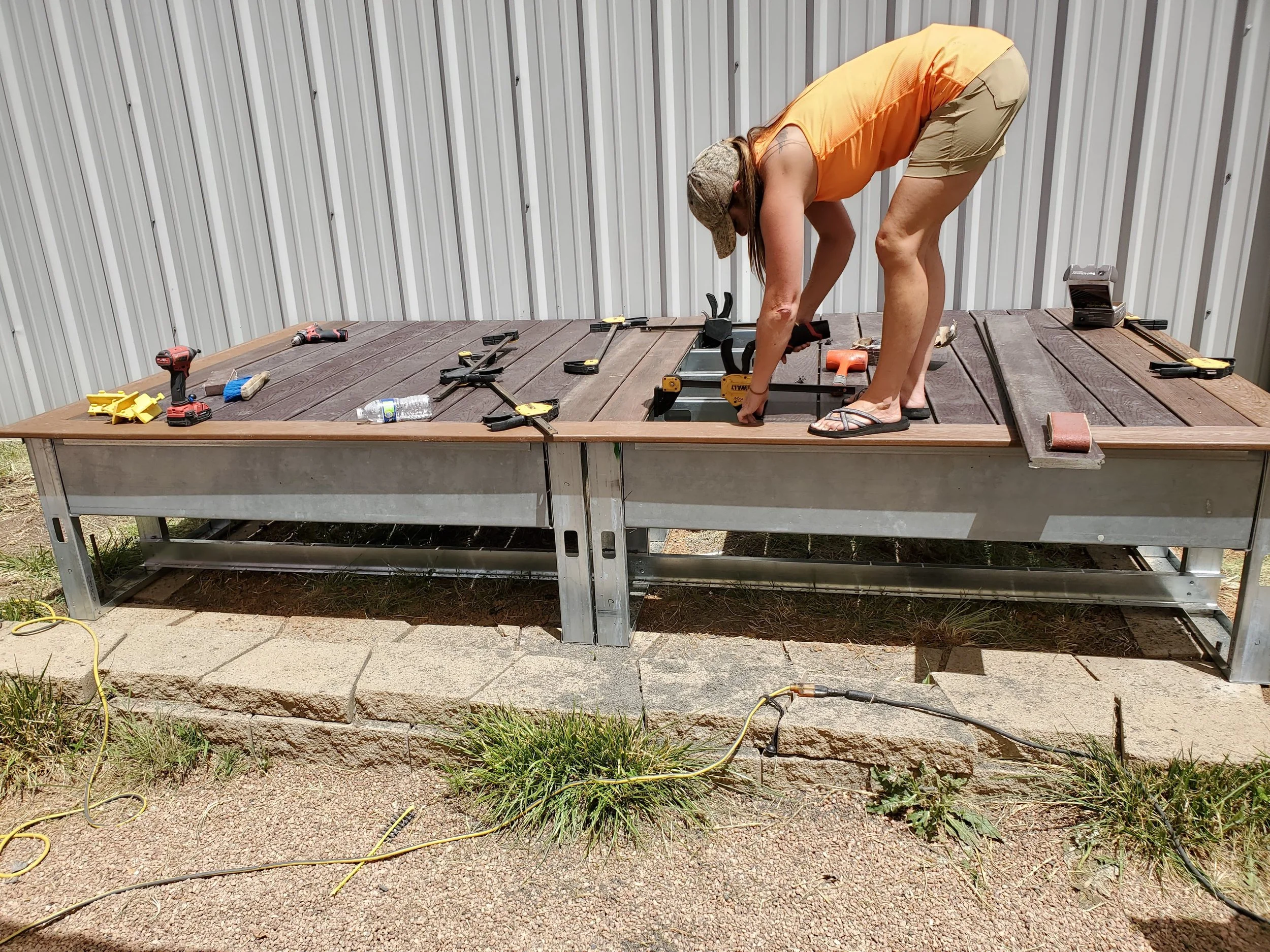 A woman working on a woodworking project outdoors, bent over a large wooden platform with tools scattered around, in front of a metal building.