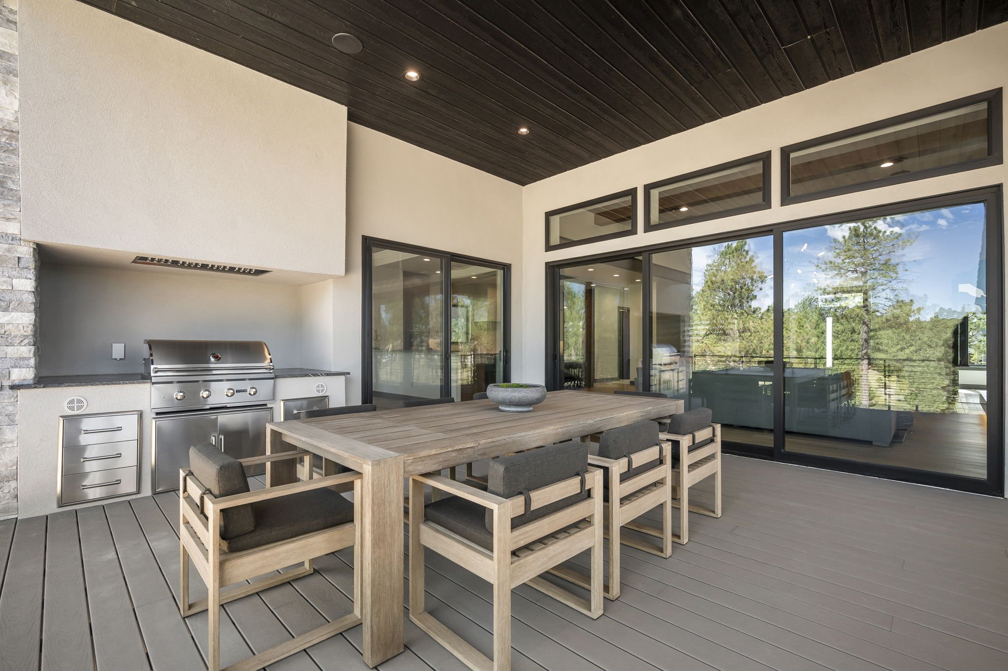 Modern outdoor patio area with a dining table, eight chairs, outdoor grill, and large glass windows overlooking a lush wooded landscape.