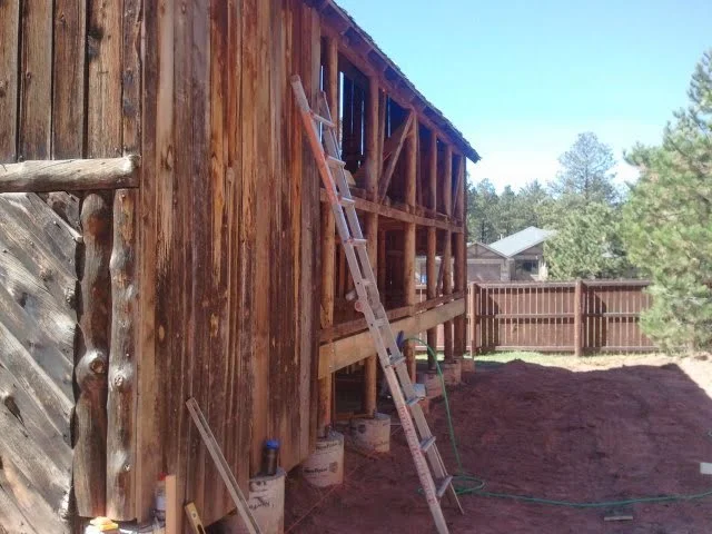Old wooden barn under repair and renovation with a ladder leaning against it surrounded by dirt and trees.