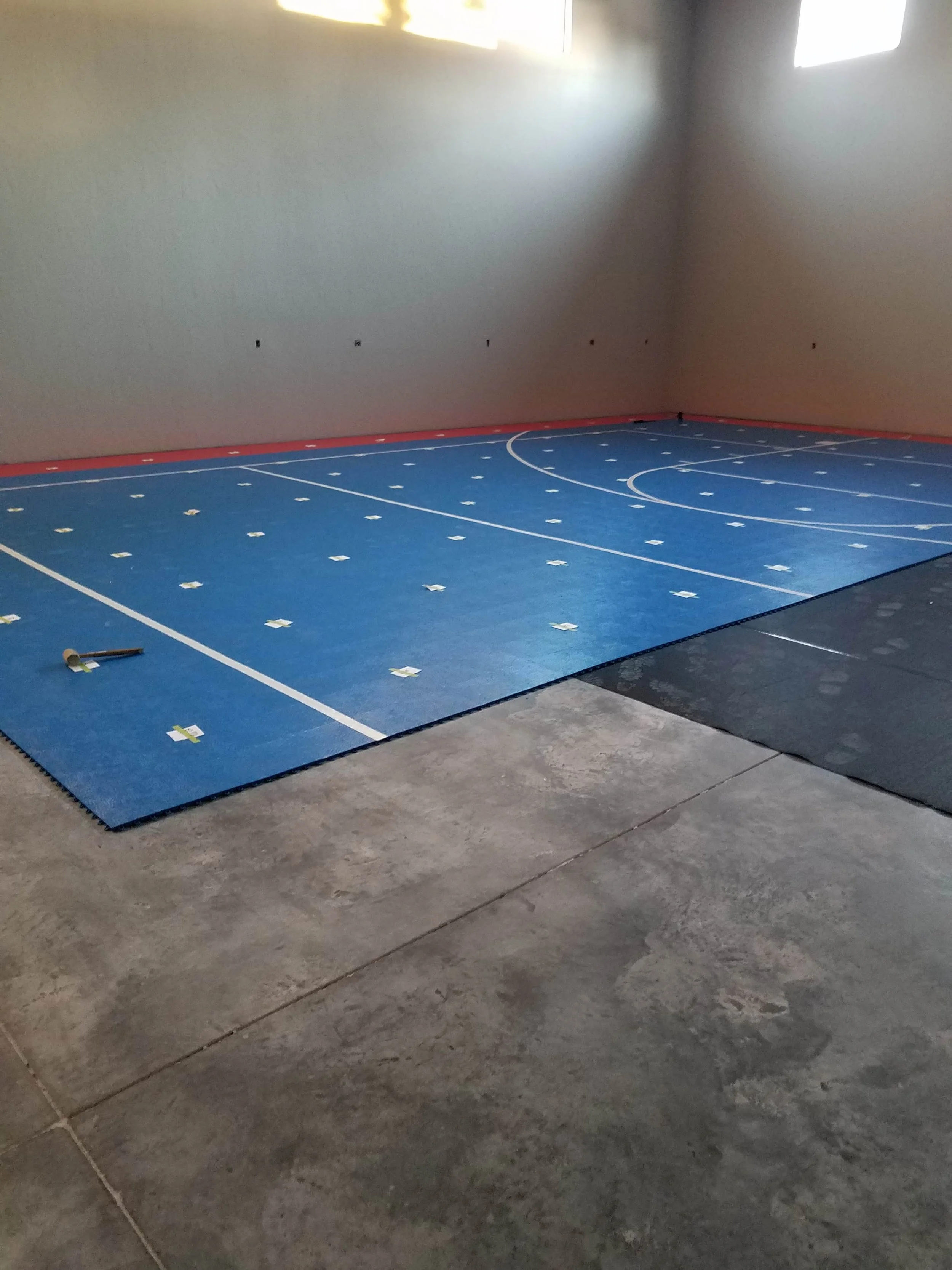 An empty basketball court with a blue floor, white lines, and orange boundary walls, located inside a RV garage with gray concrete floors and walls. There are protective black mats on the foreground and two small windows near the ceiling.
