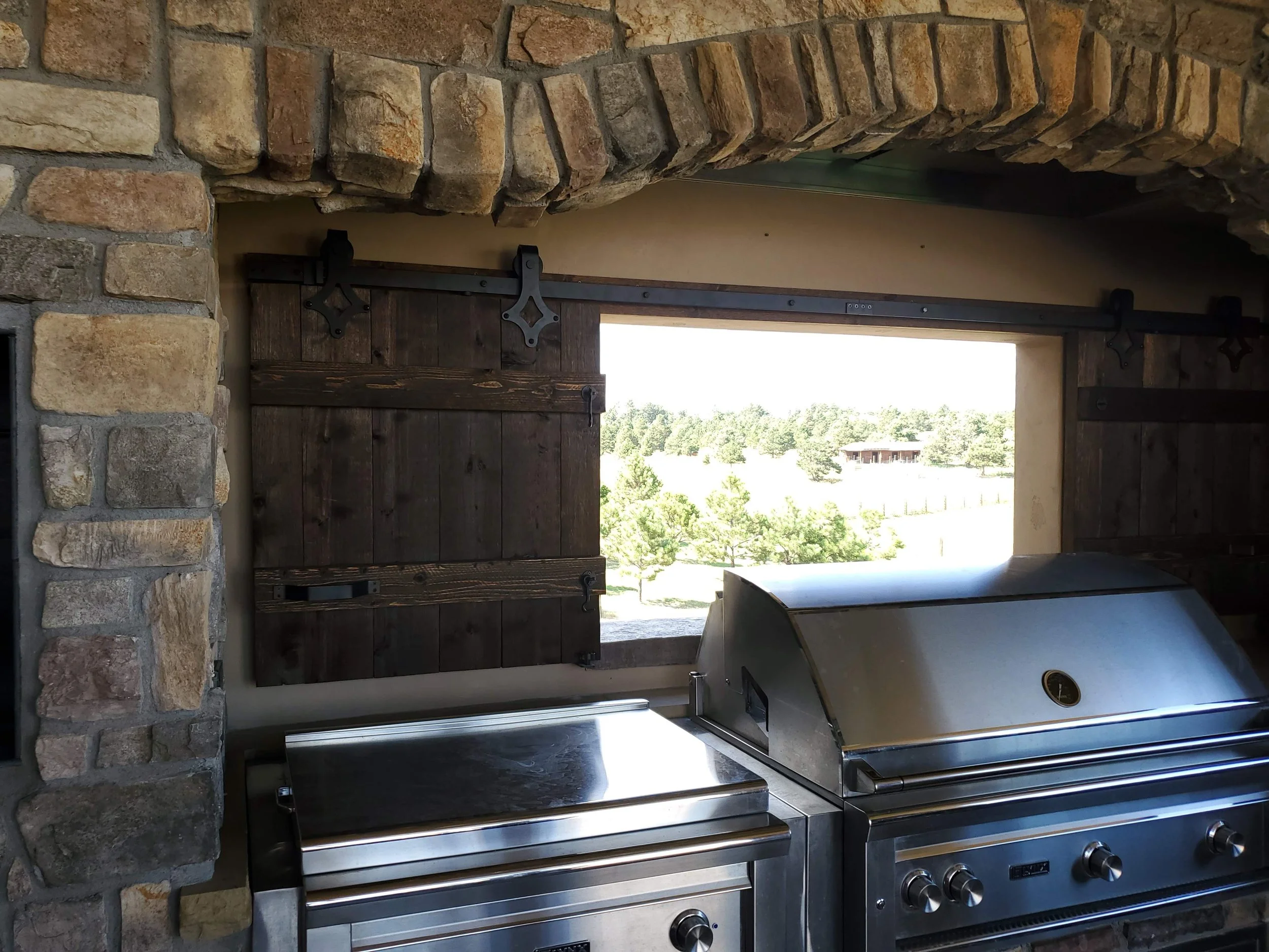 Stainless steel outdoor grill with four burners and temperature dials, set against a stone wall with a window showing trees and a house in the background.