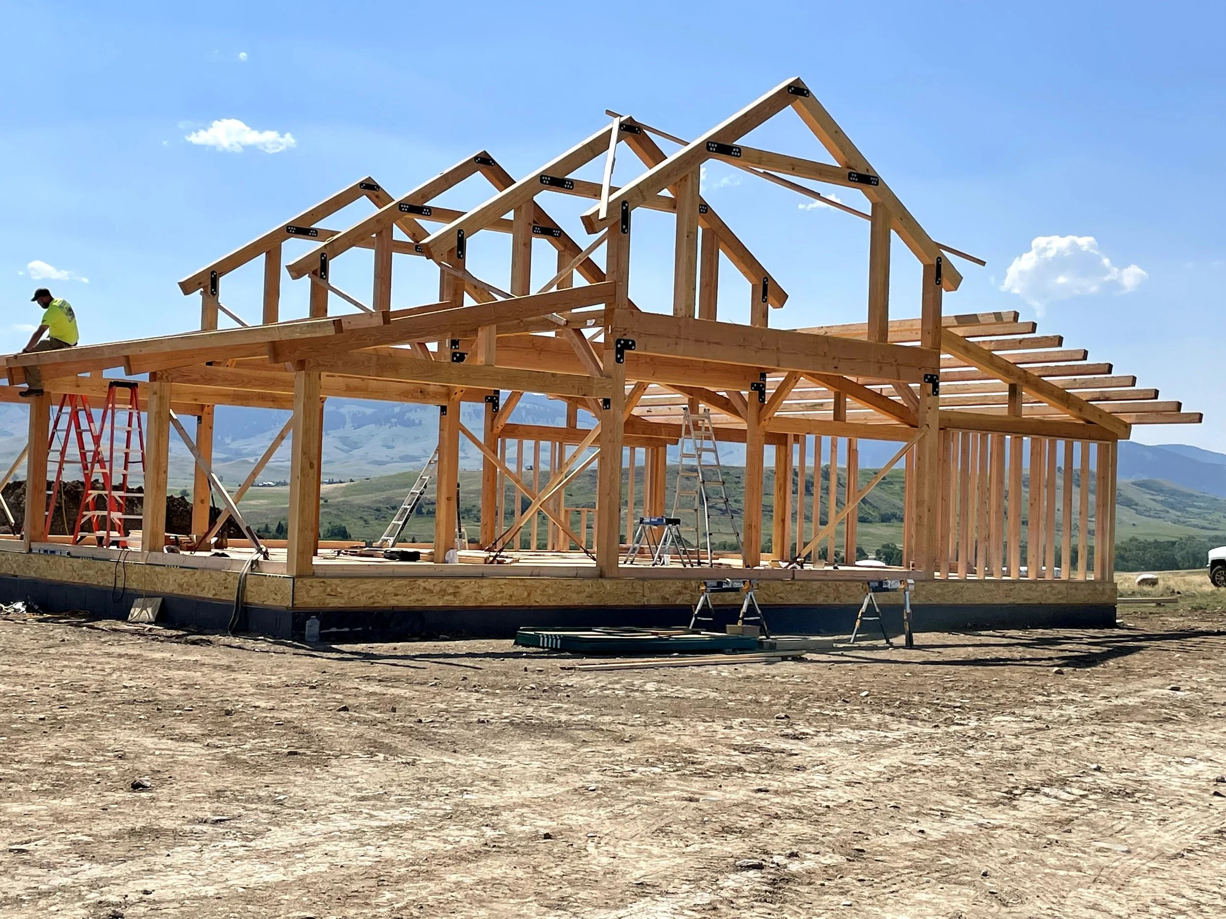 Wooden framing of a Post and Beam house under construction with ladders, construction workers, and tools, in a rural area with mountains in the background.