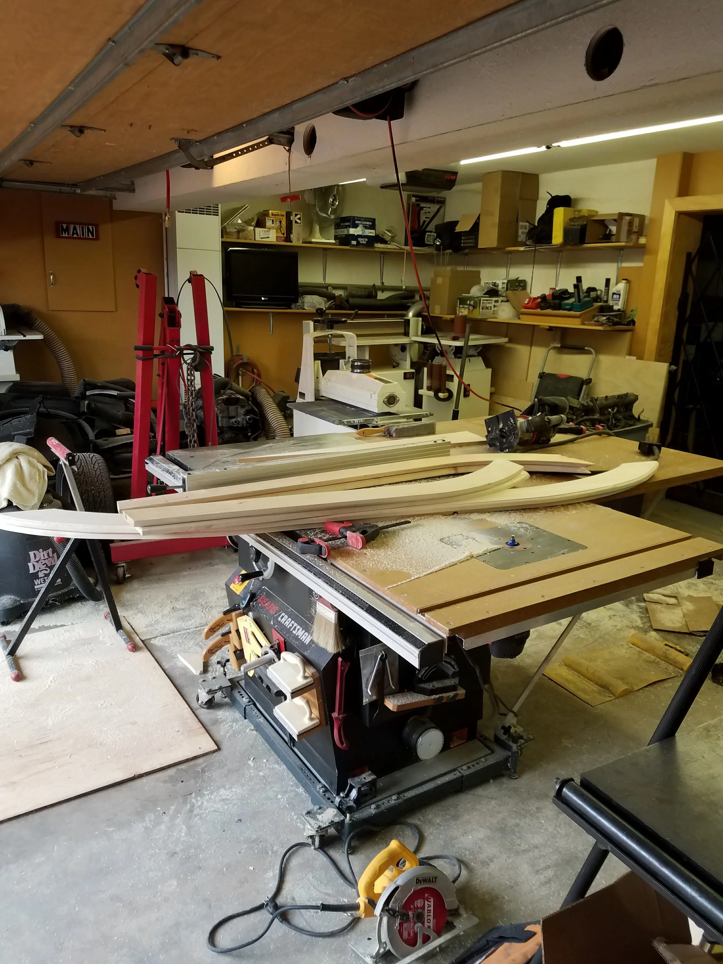 A woodworking workshop with a table saw, various woodworking tools, wooden pieces, and a cluttered workbench area.