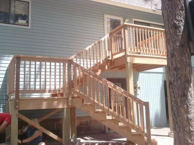 Newly built wooden outdoor staircase and deck attached to a house with siding, surrounded by trees.