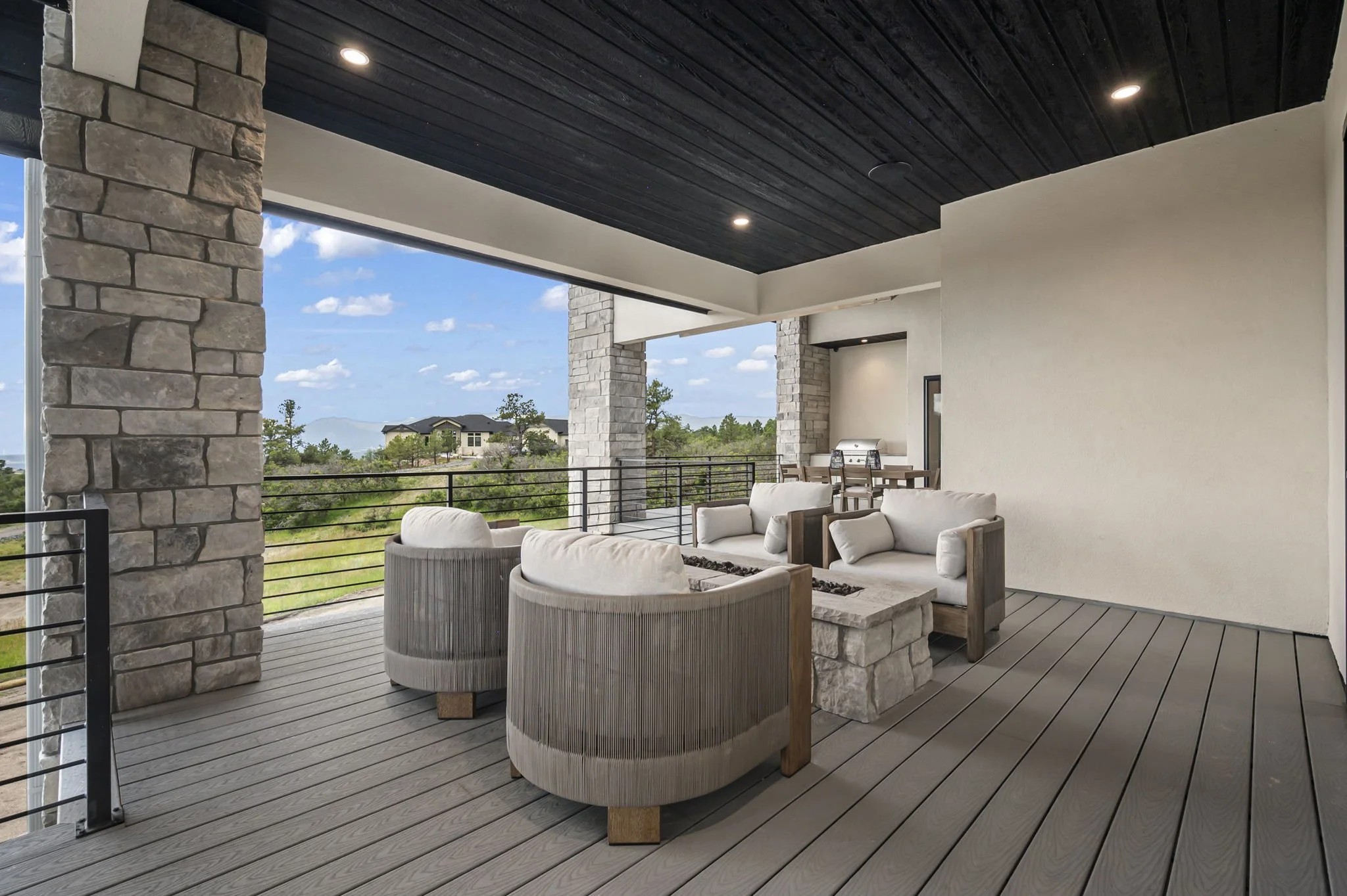 Outdoor covered patio with stone pillars, wooden ceiling, and furniture including cushioned chairs, a sofa, and a dining table, with a view of distant houses and trees.