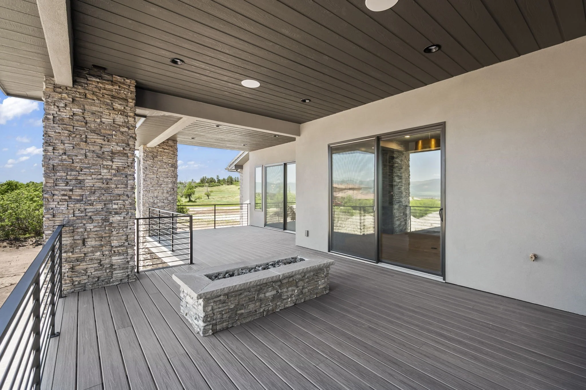 Modern outdoor balcony with stone columns, sliding glass doors, wood-paneled ceiling with recessed lights, concrete planter, and view of a green landscape.