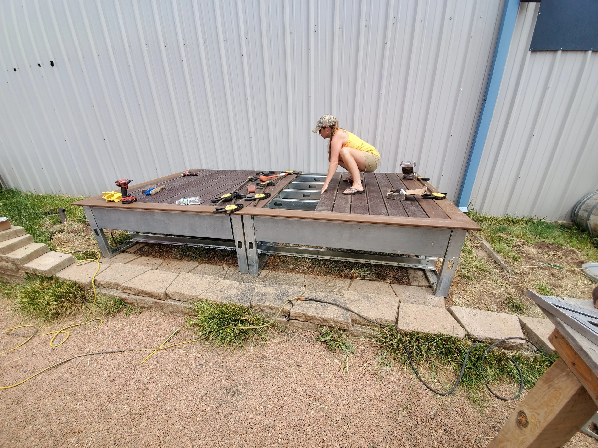 A woman in a yellow tank top, shorts, and a camouflage cap squats on a wooden deck with tools scattered around, working on an outdoor project near a large metal building.