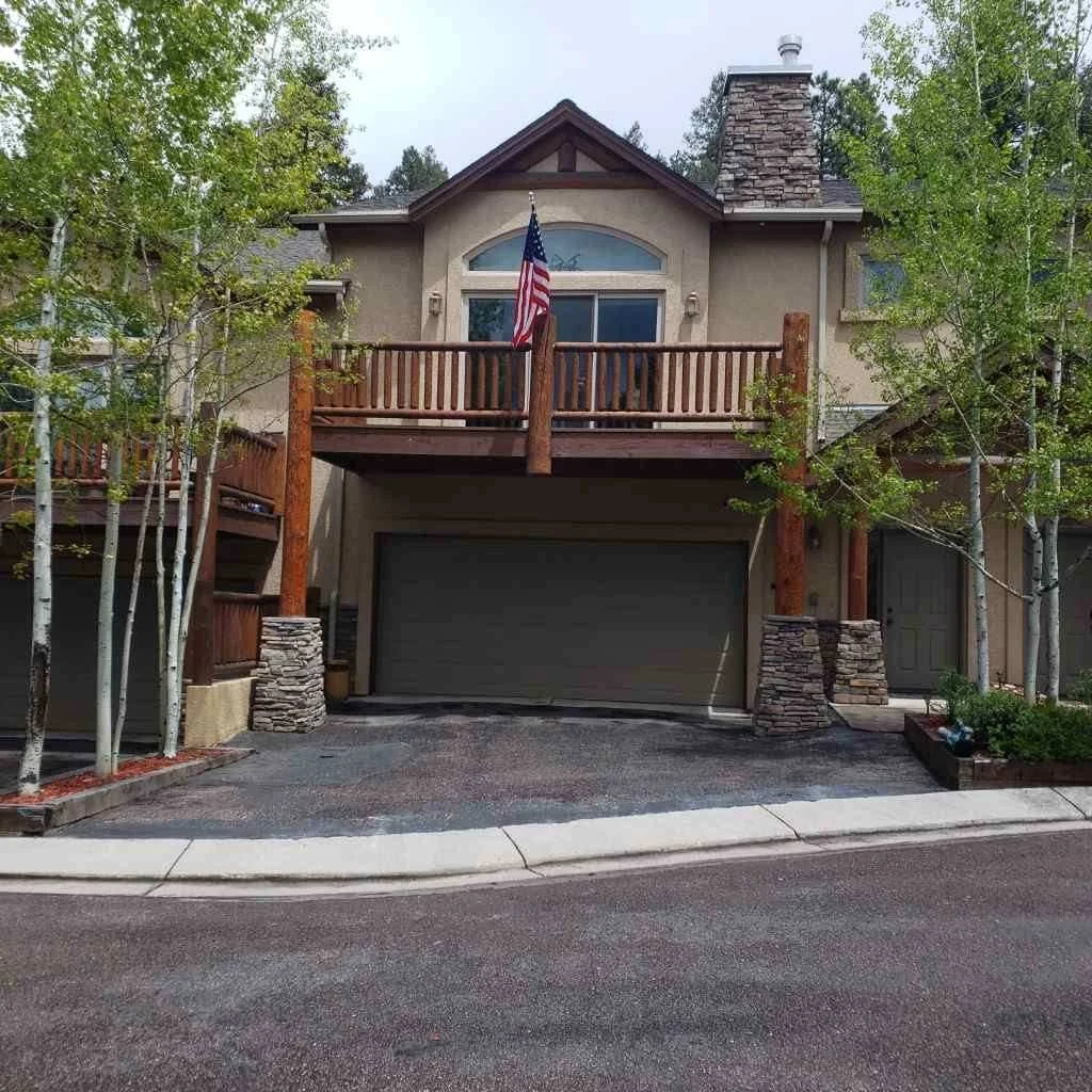 Front view of a two-story house with a garage, a wooden balcony with an American flag, surrounded by trees and landscaping.