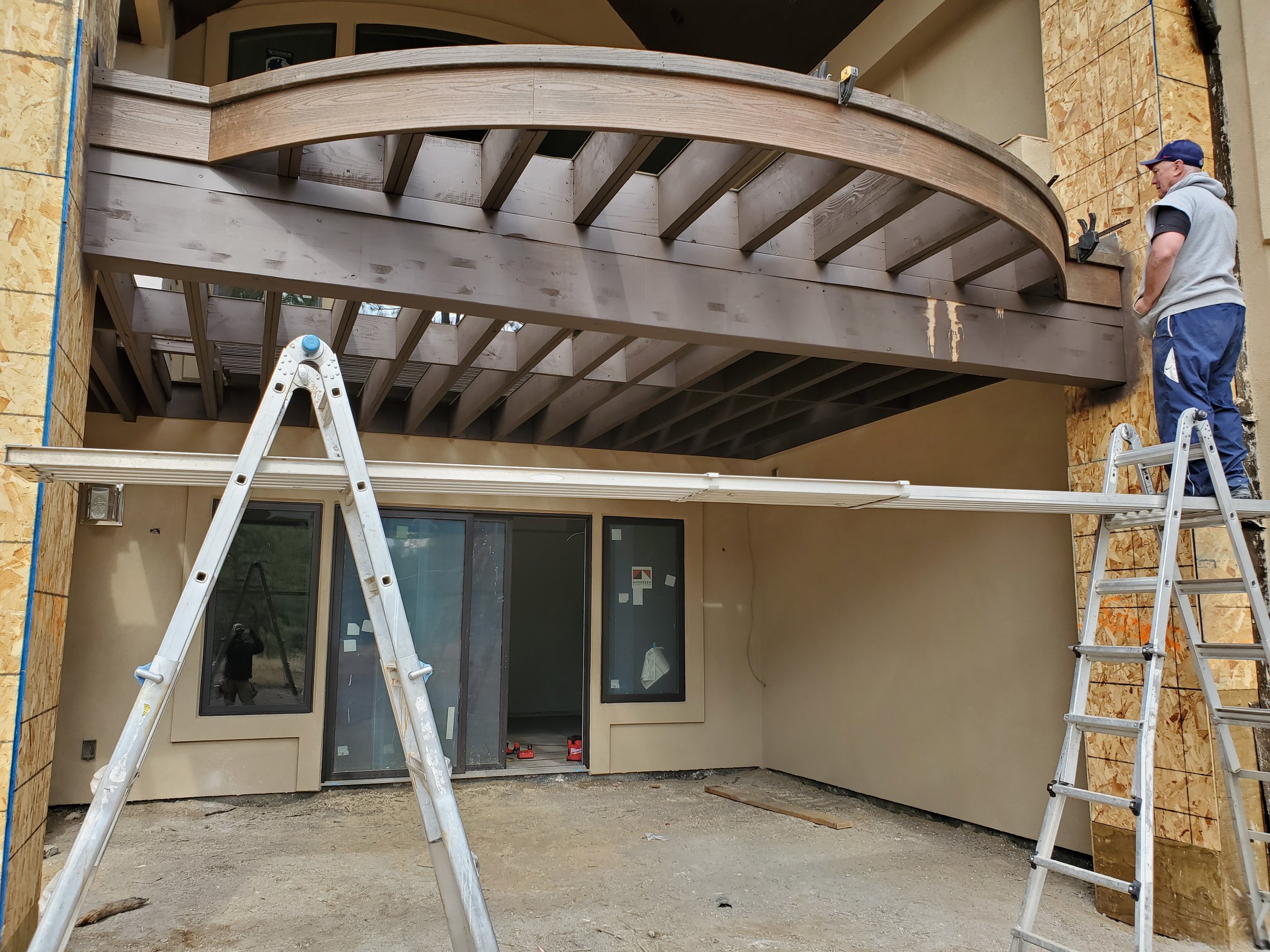 Construction site of a curved deck with wood joist framing , two ladders, and a man working on a scaffold board it, stucco walls unfinished stone columns