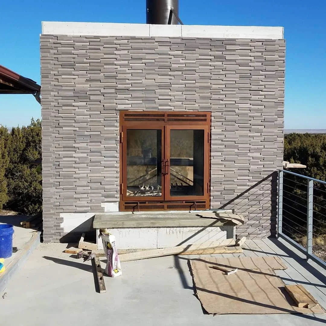 A construction site featuring a modern building with a textured gray brick exterior, a wooden framed sliding glass door, and a concrete deck in progress. Tools and materials are scattered on the concrete surface, with a railing on the right side, ove