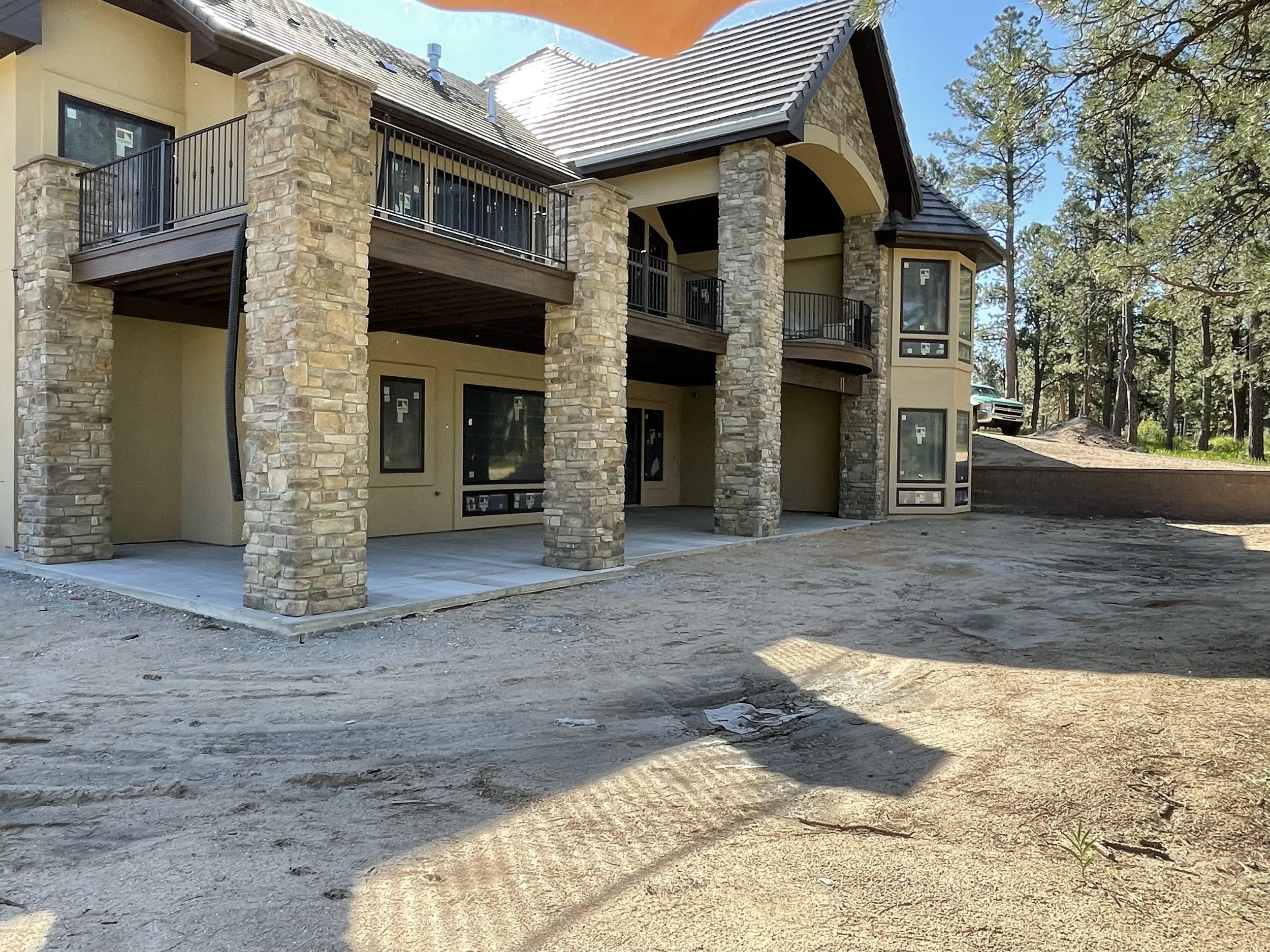 A multi-story house under construction with stone pillars and beige walls. The house features large windows and a balcony with black railings, situated in a wooded area with trees and a dirt ground.