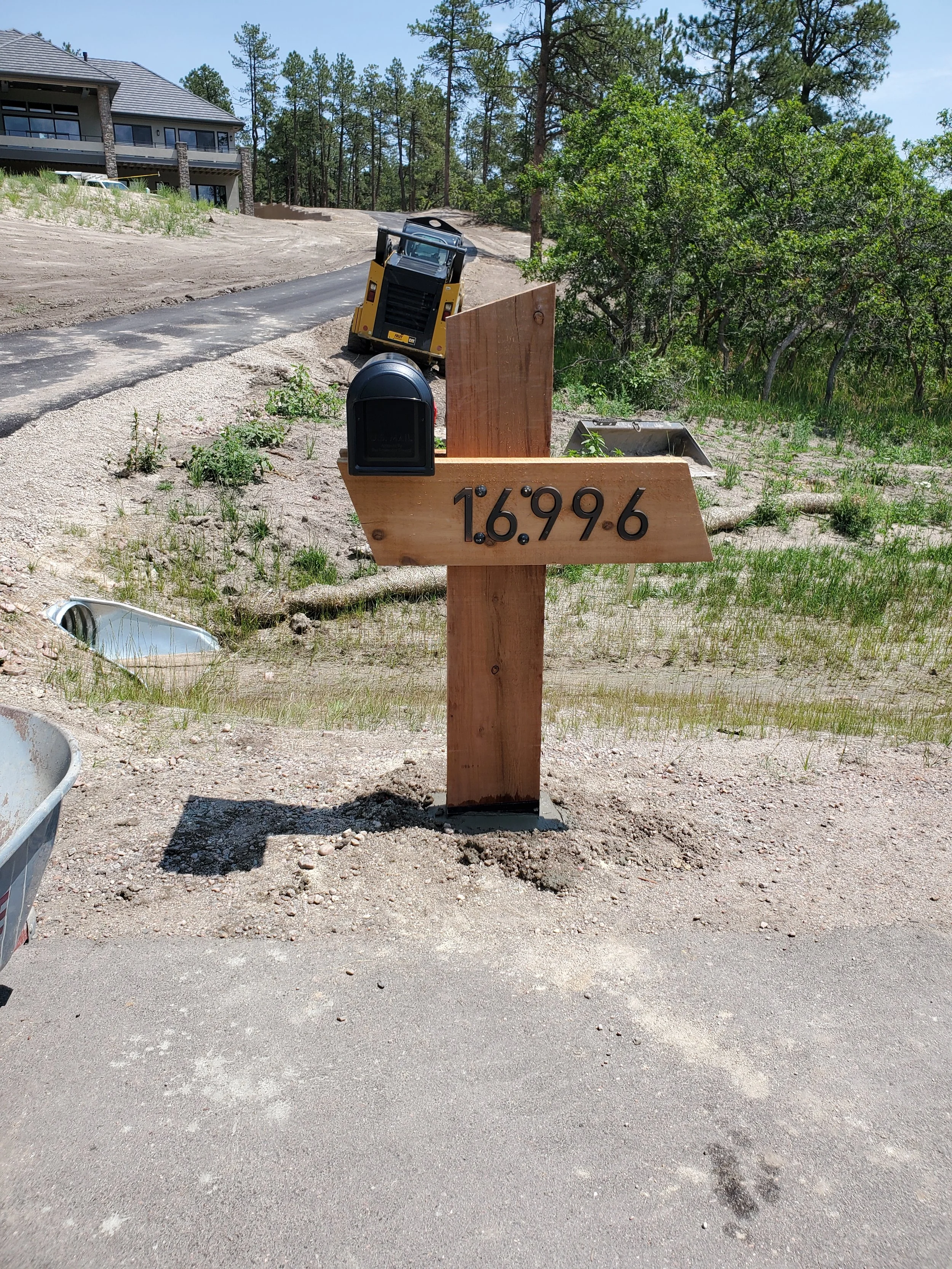 A wooden sign with the coordinates 16.996 is placed in a construction area with a small yellow construction vehicle in the background, dirt road, and a house on the hill surrounded by trees.
