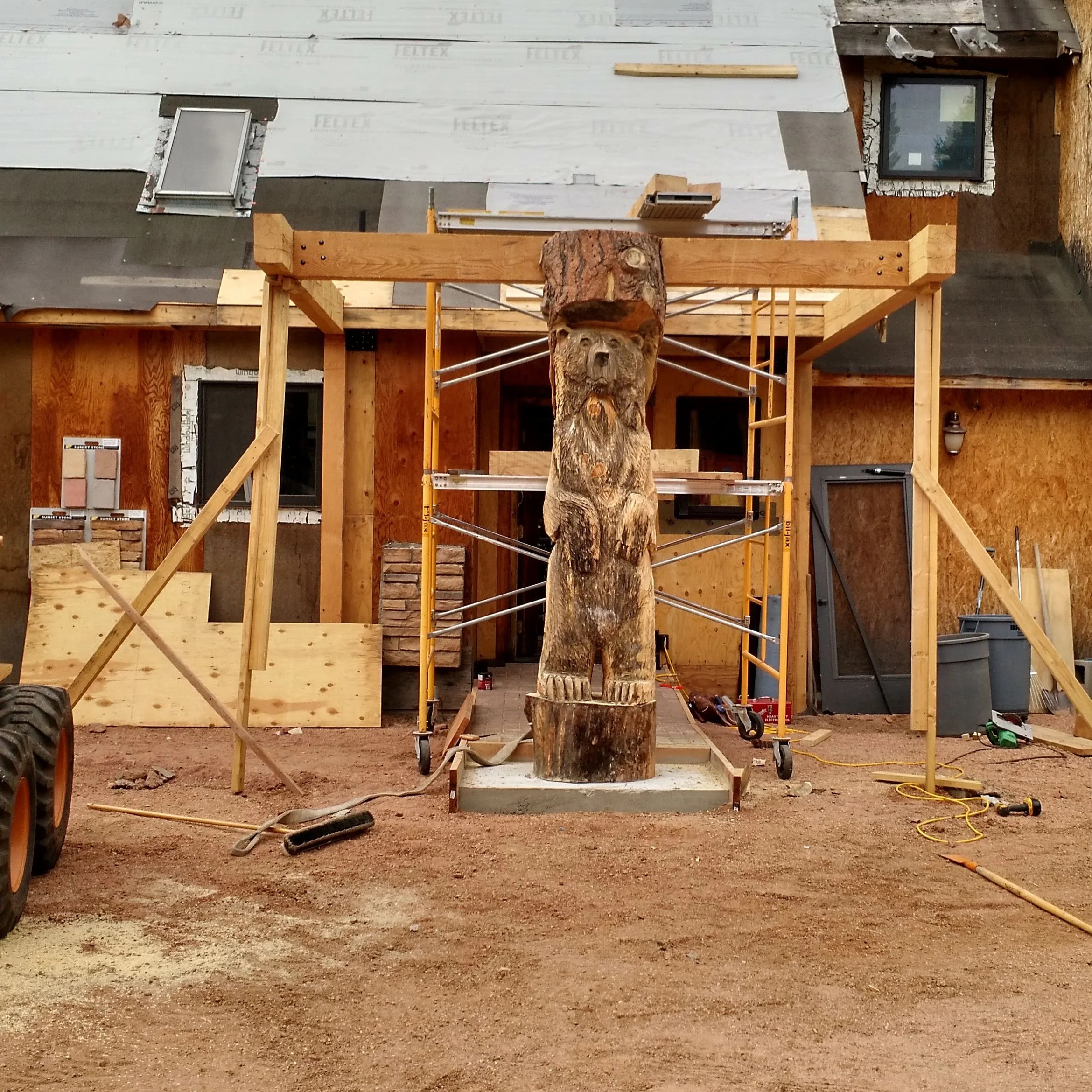 Construction site with wooden framing, a log in the center, and a building under construction with windows and insulation. The ground is bare dirt, and construction tools and materials are scattered around.