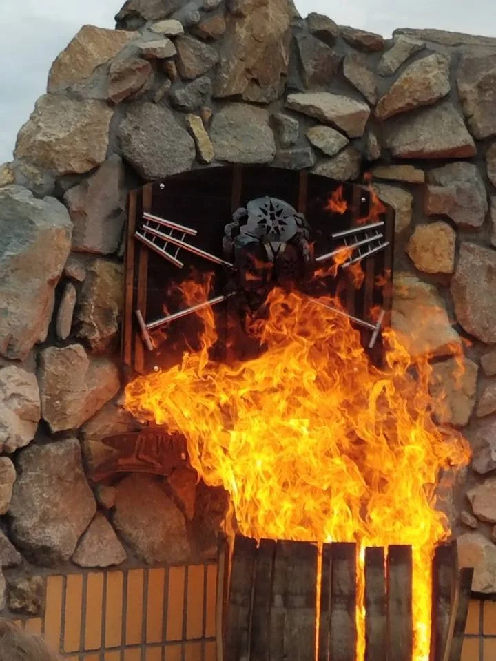 A large fire burning inside a metal container, placed against a stone wall. Behind the fire, there is a decorative wall-mounted iron piece in the shape of a skull with crossed rifles.